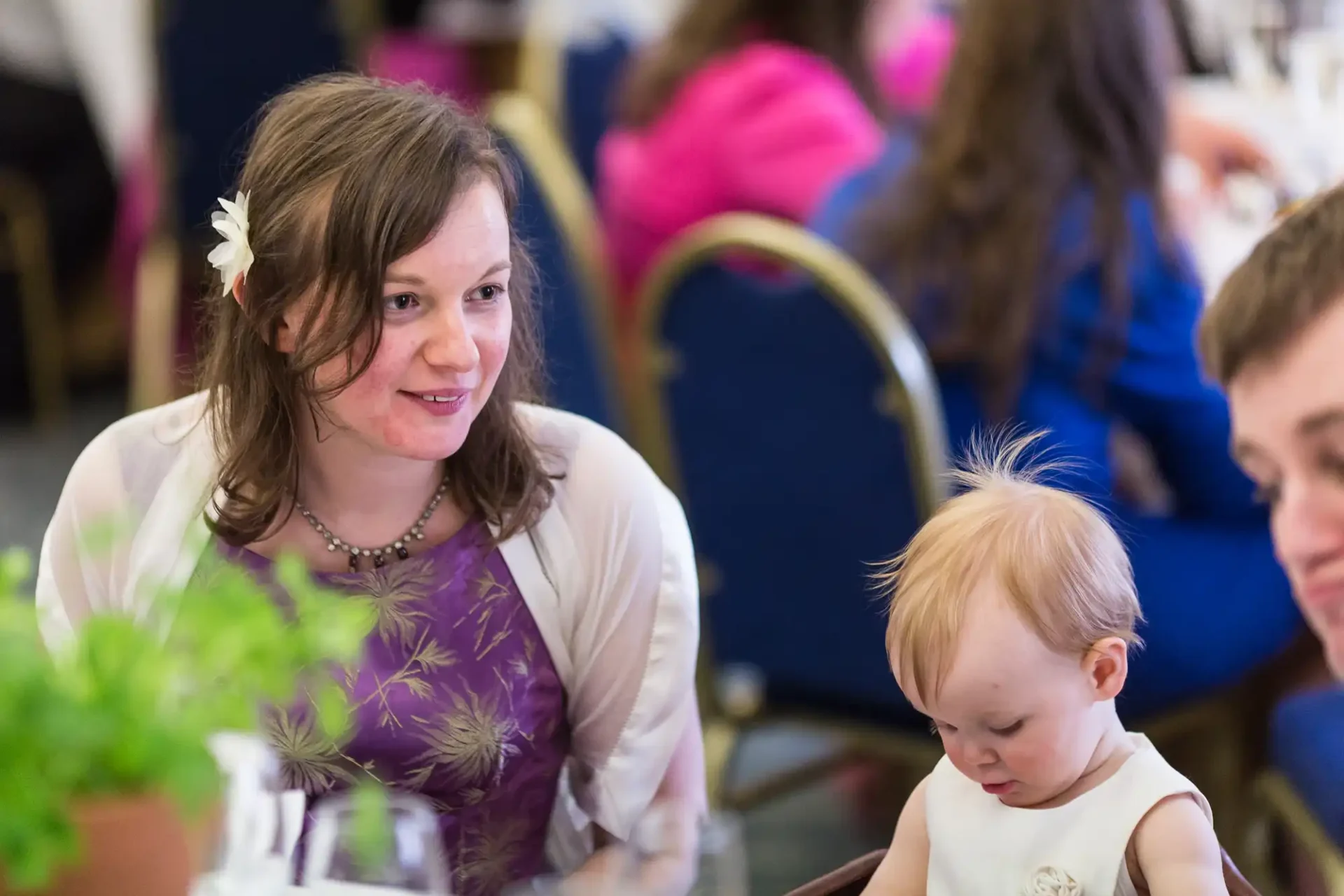 Woman in floral dress smiling at baby in white outfit at a formal event.