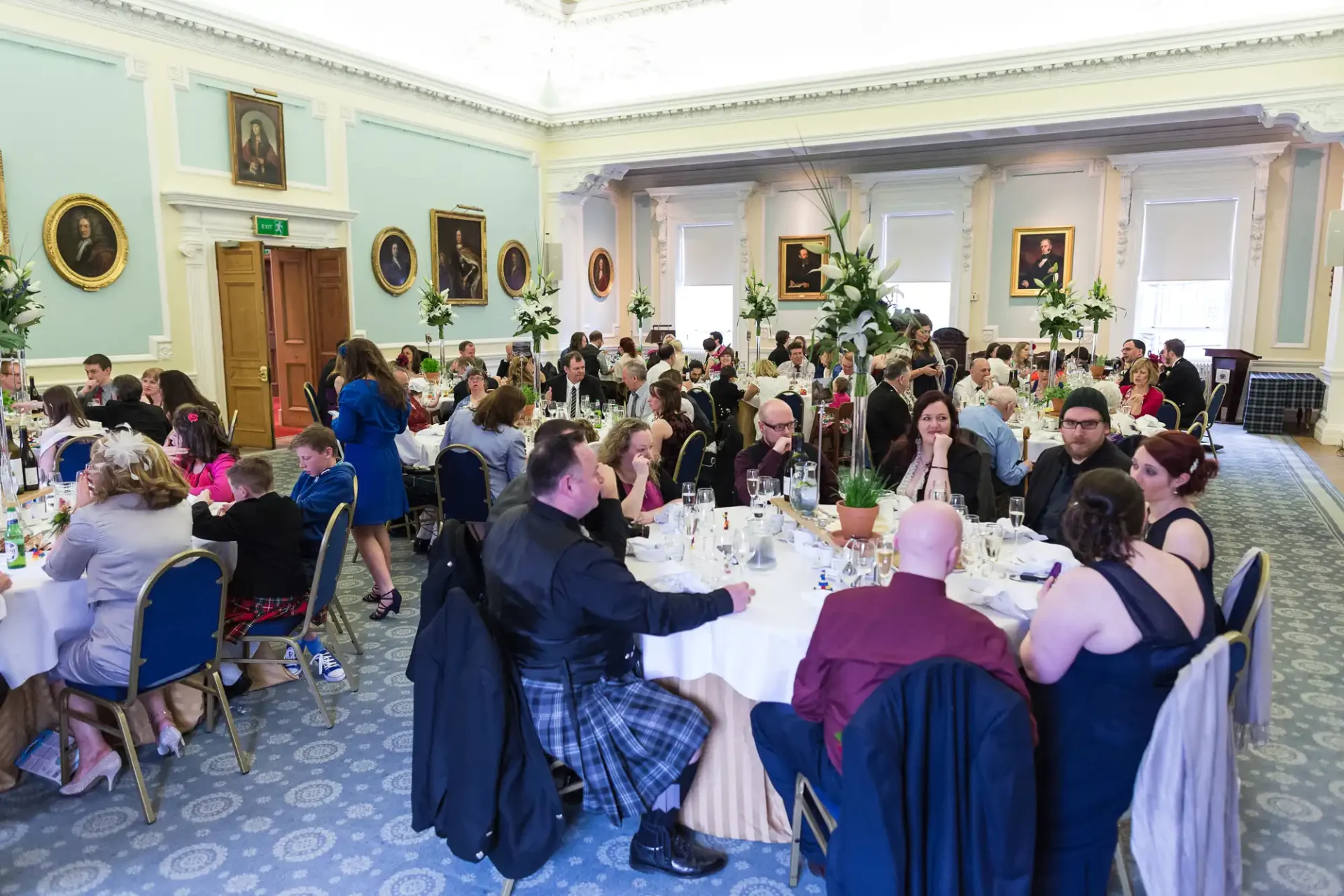 Guests in formal attire dine at round tables in an elegant room with chandeliers and portraits on the walls.
