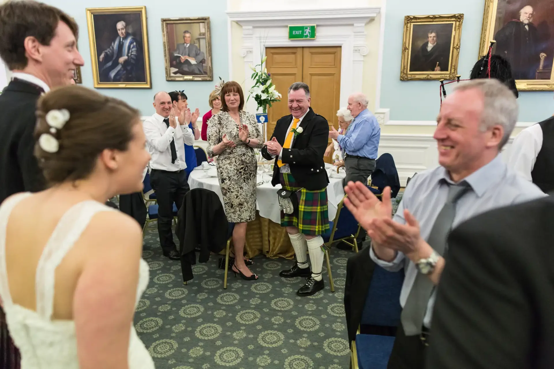 Wedding guests clapping and smiling at the bride as she enters the room, decorated with historical portraits, at a reception.