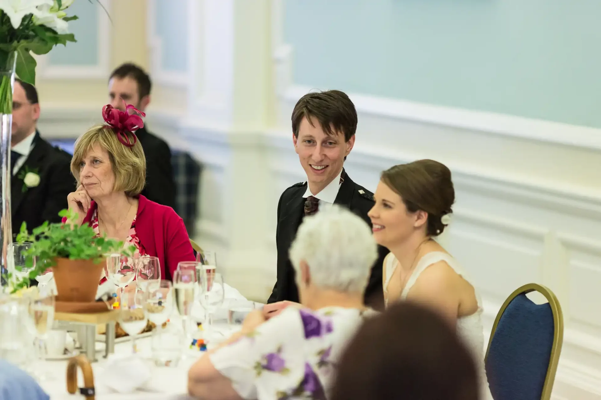 Guests at a wedding reception, including a bride and another person smiling, seated at a table with several attendees and dining sets in a brightly lit room.
