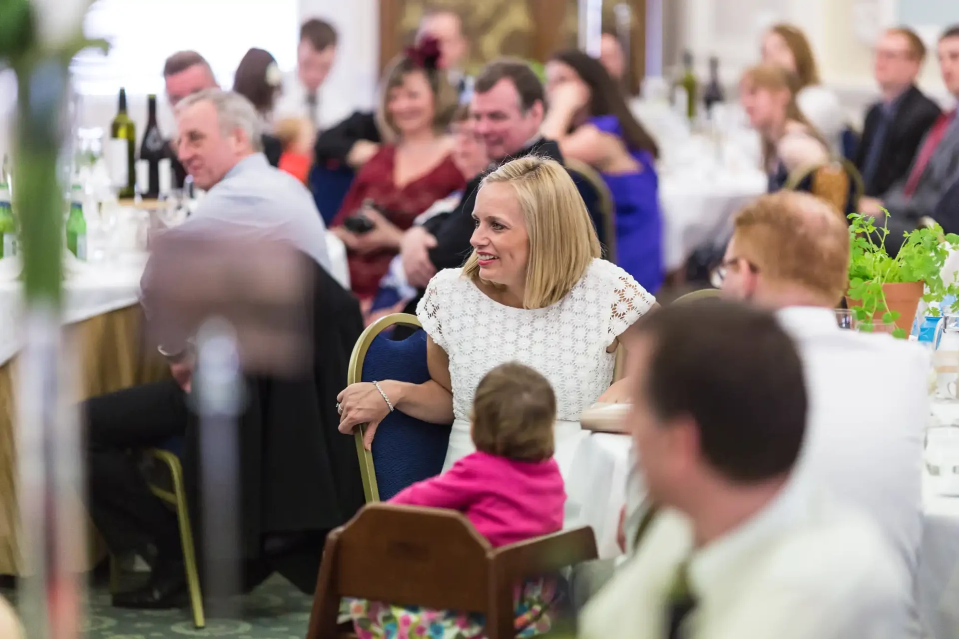 A woman in a white dress smiles while seated at a crowded event, with guests at tables engaging in conversations around her.