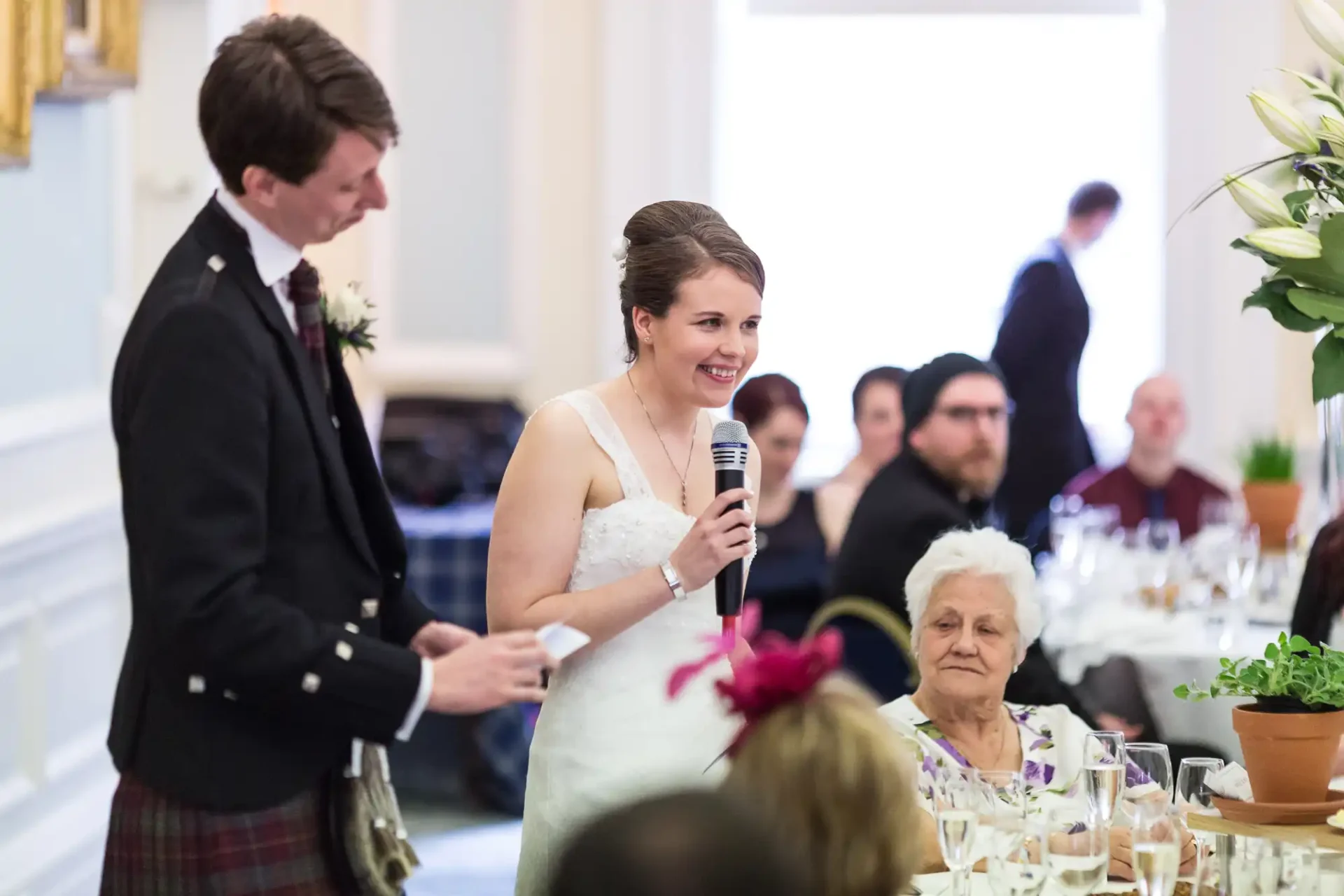 A bride speaking into a microphone at a wedding reception, with a groom and elderly woman seated nearby, in an elegant dining room.