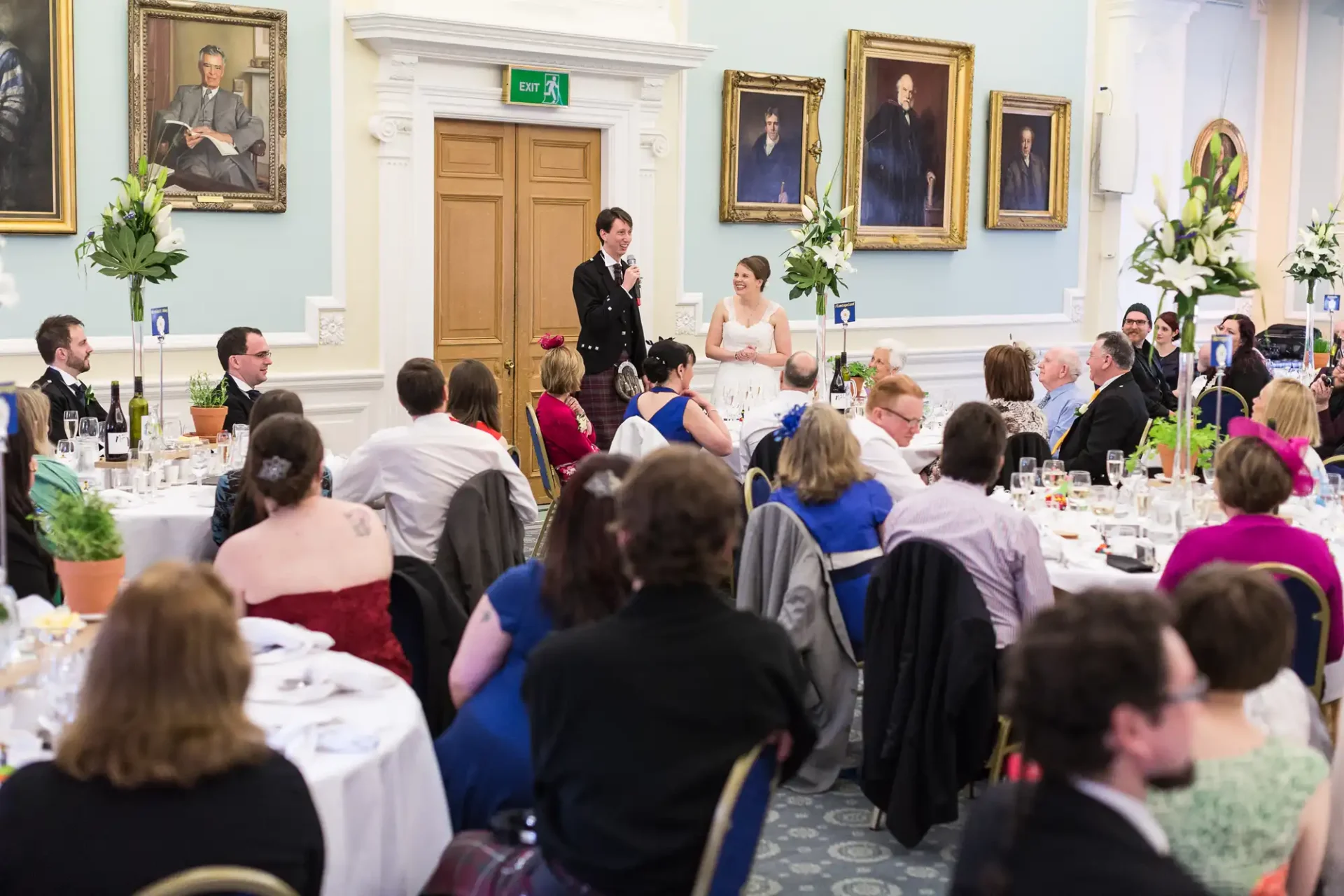Bride and groom standing together in a banquet hall filled with seated guests during a wedding reception, with portraits on the walls.