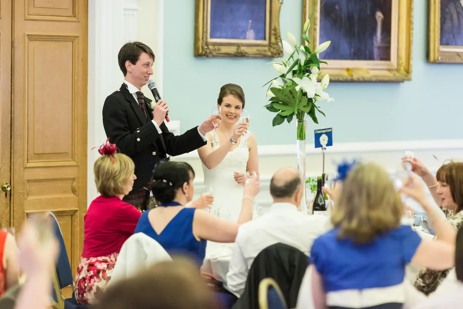 A bride and groom giving a speech at their wedding reception, surrounded by seated guests in a decorated hall.