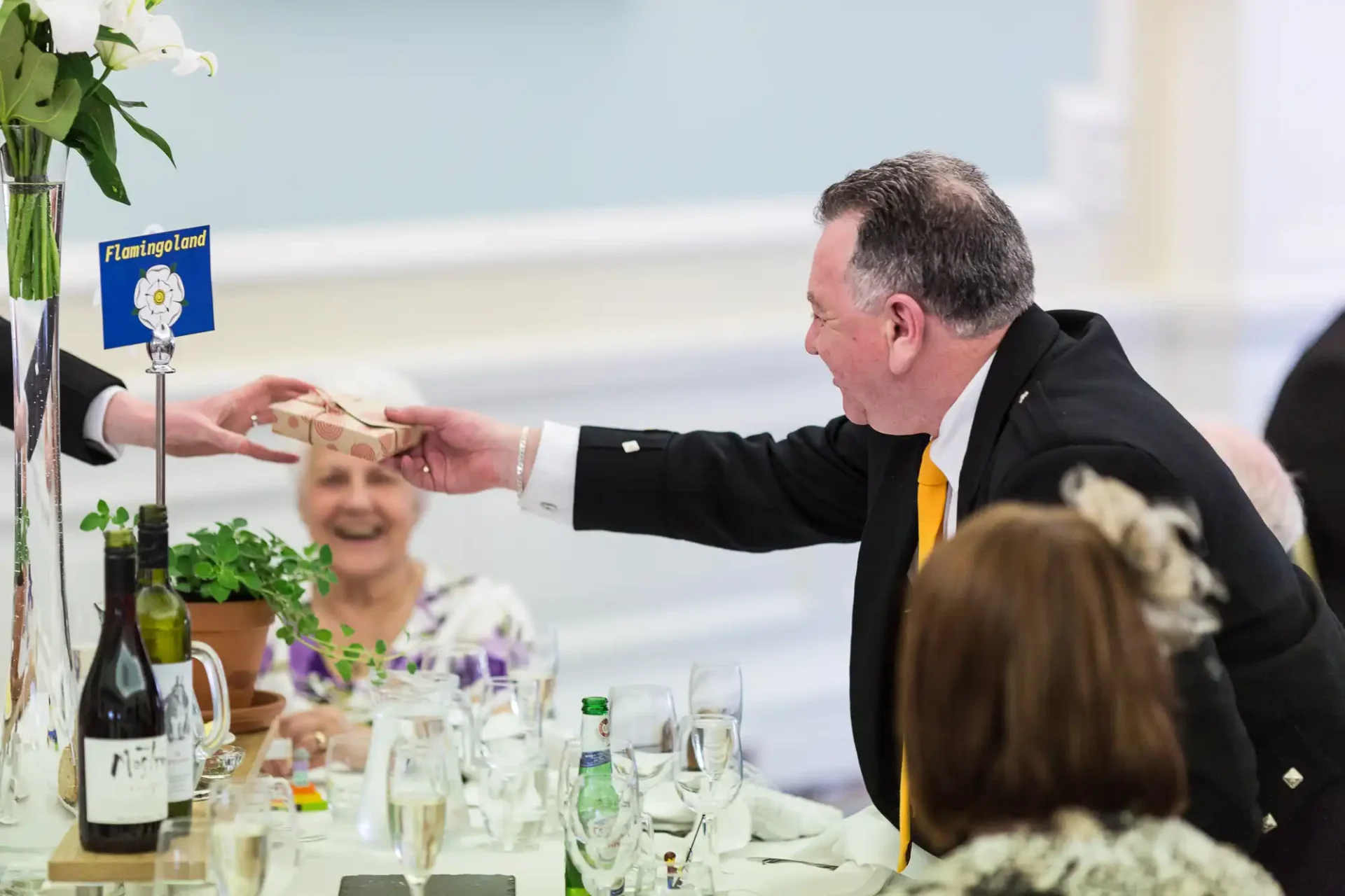 A man in a black suit passes a wooden doll across a dining table to a smiling woman, as others look on in a brightly lit room.