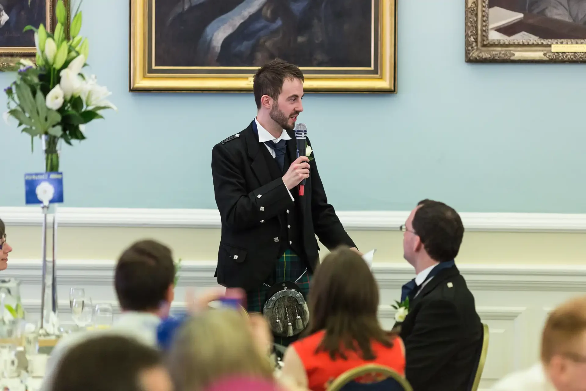 A man in a formal uniform speaks into a microphone at a dinner event, addressing seated guests in a decorated room with paintings.