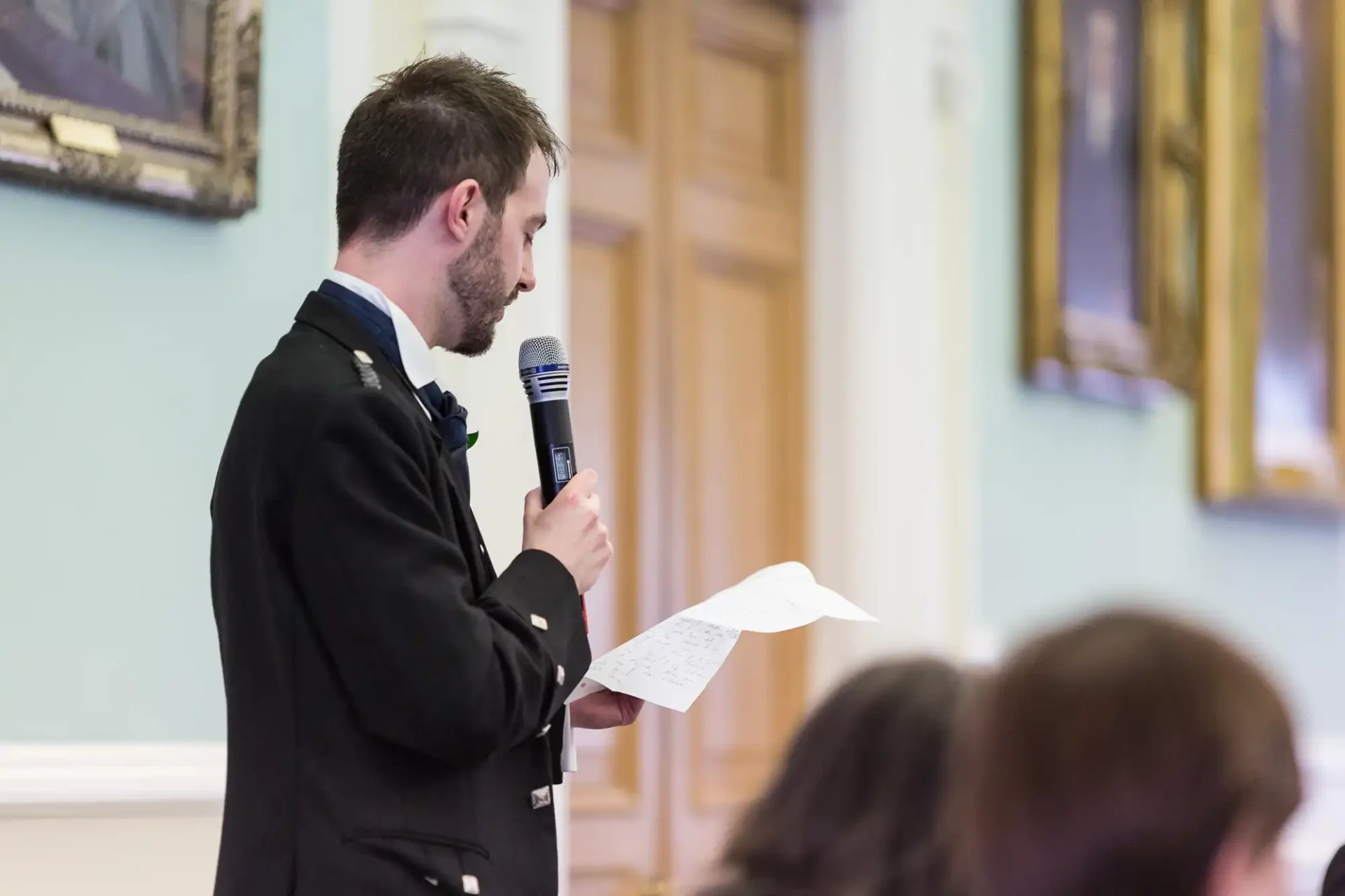Man in formal attire speaking into a microphone while holding papers, standing in a room adorned with paintings.