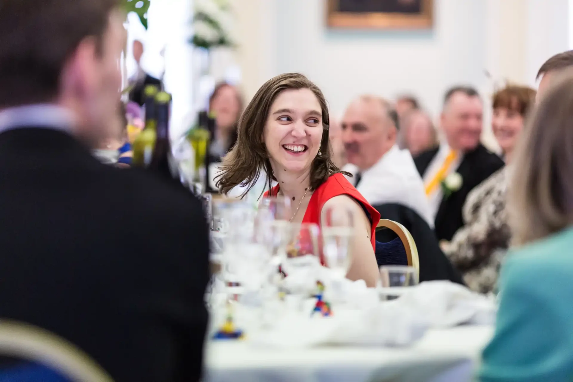 A smiling woman in a formal setting with other guests, seated at a dinner table adorned with glasses and decor.