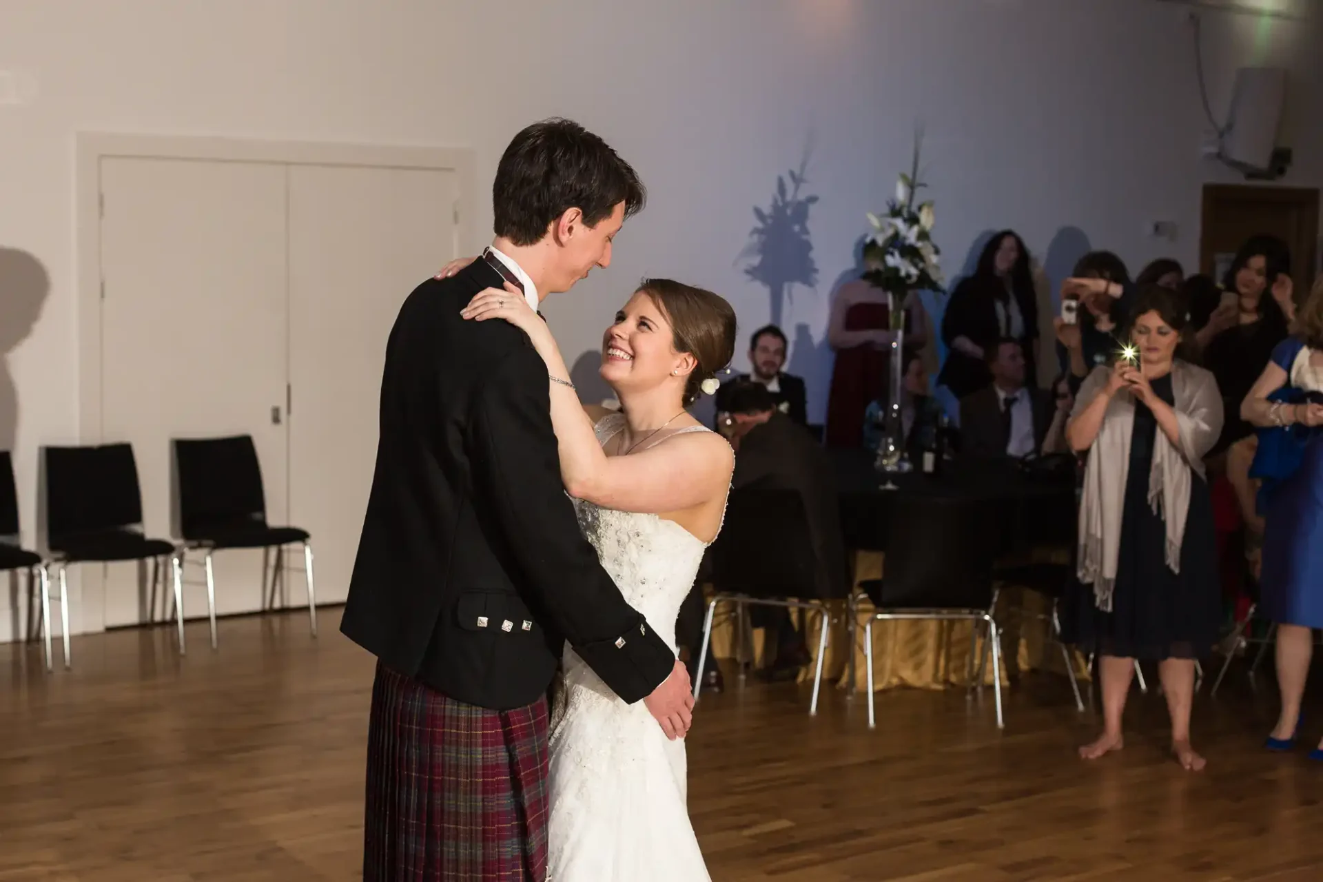 A bride and groom share a dance at their wedding reception, surrounded by guests in a well-lit hall.
