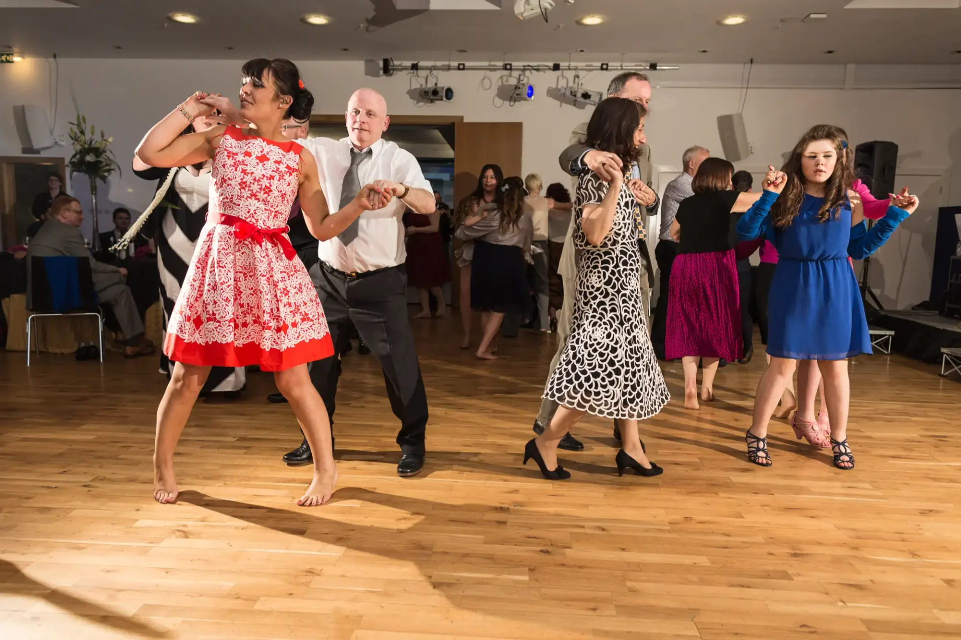Four people of varying ages dance together in a lively ballroom, two women in colorful dresses lead their male partners, with onlookers in the background.