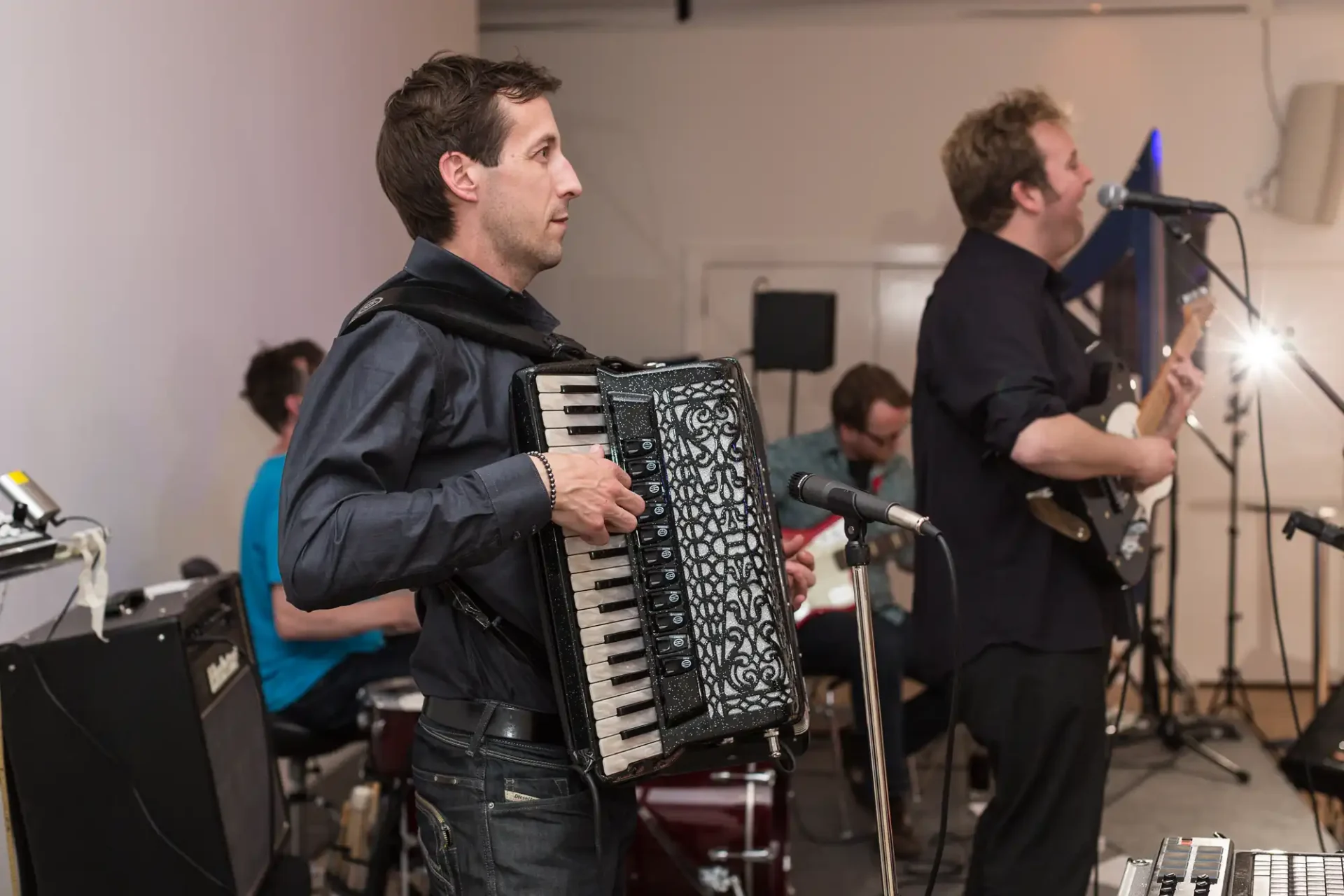 A man playing an accordion on stage with band members in the background, including a guitarist and a drummer.