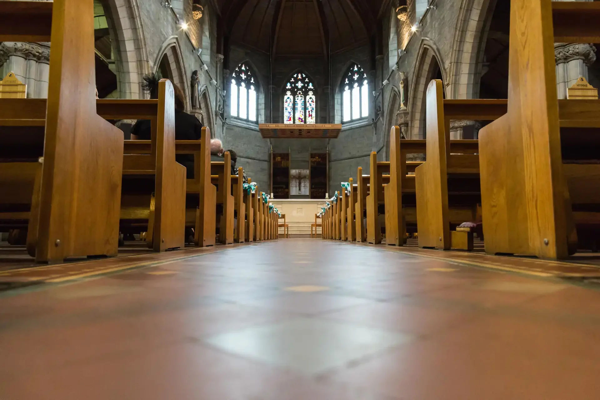 Low angle view inside a church showing rows of wooden pews leading towards an altar with stained glass windows above.