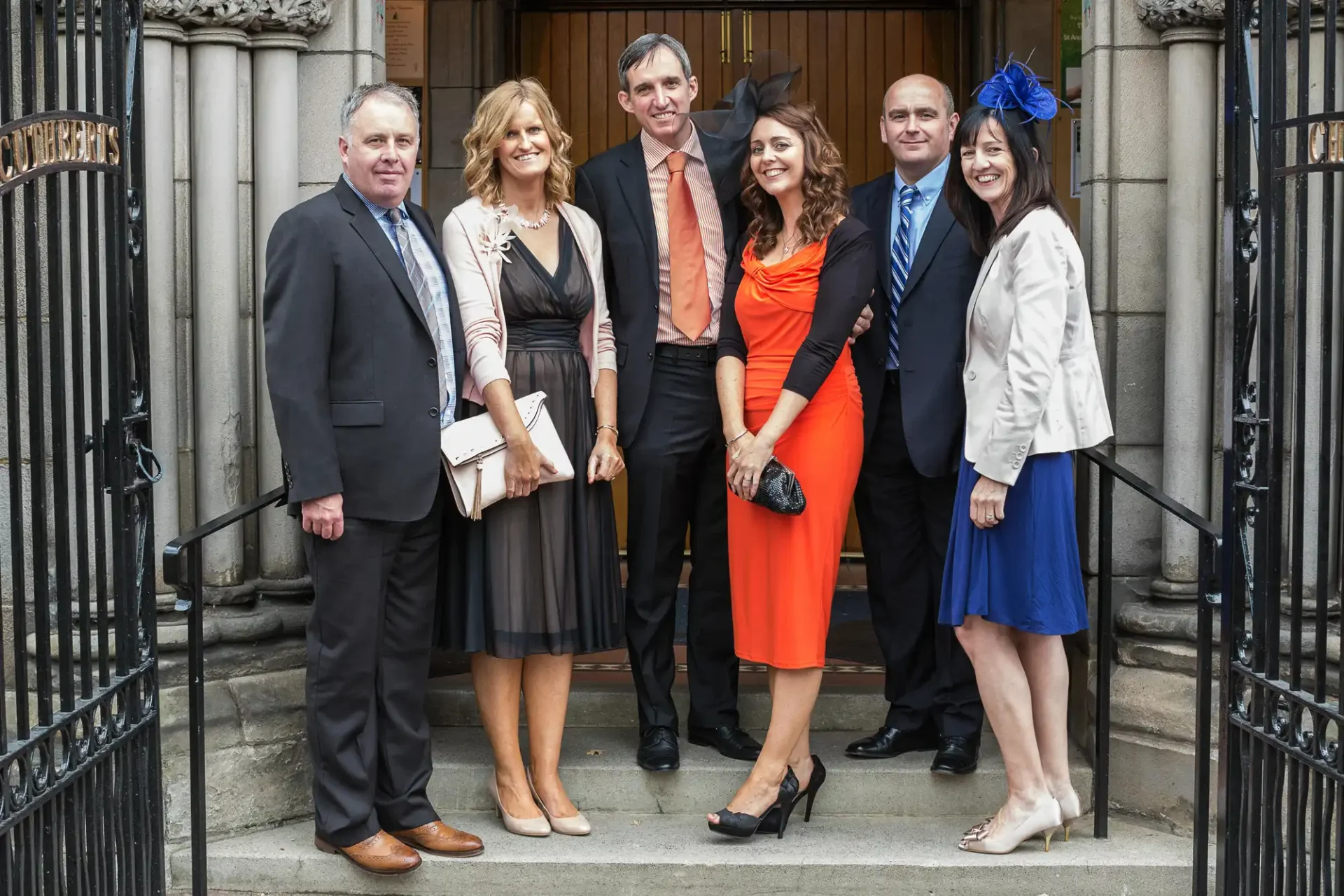 Three men and three women dressed in smart attire, posing together on steps outside a building with ornate iron railings.