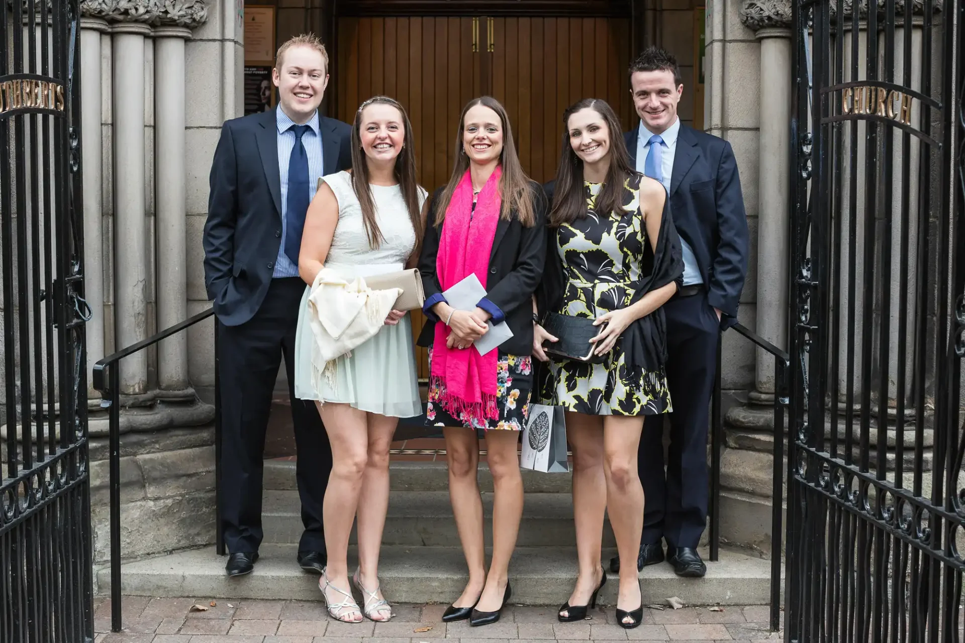 Five young adults smiling and posing together at a gate, dressed in formal attire for a special occasion.