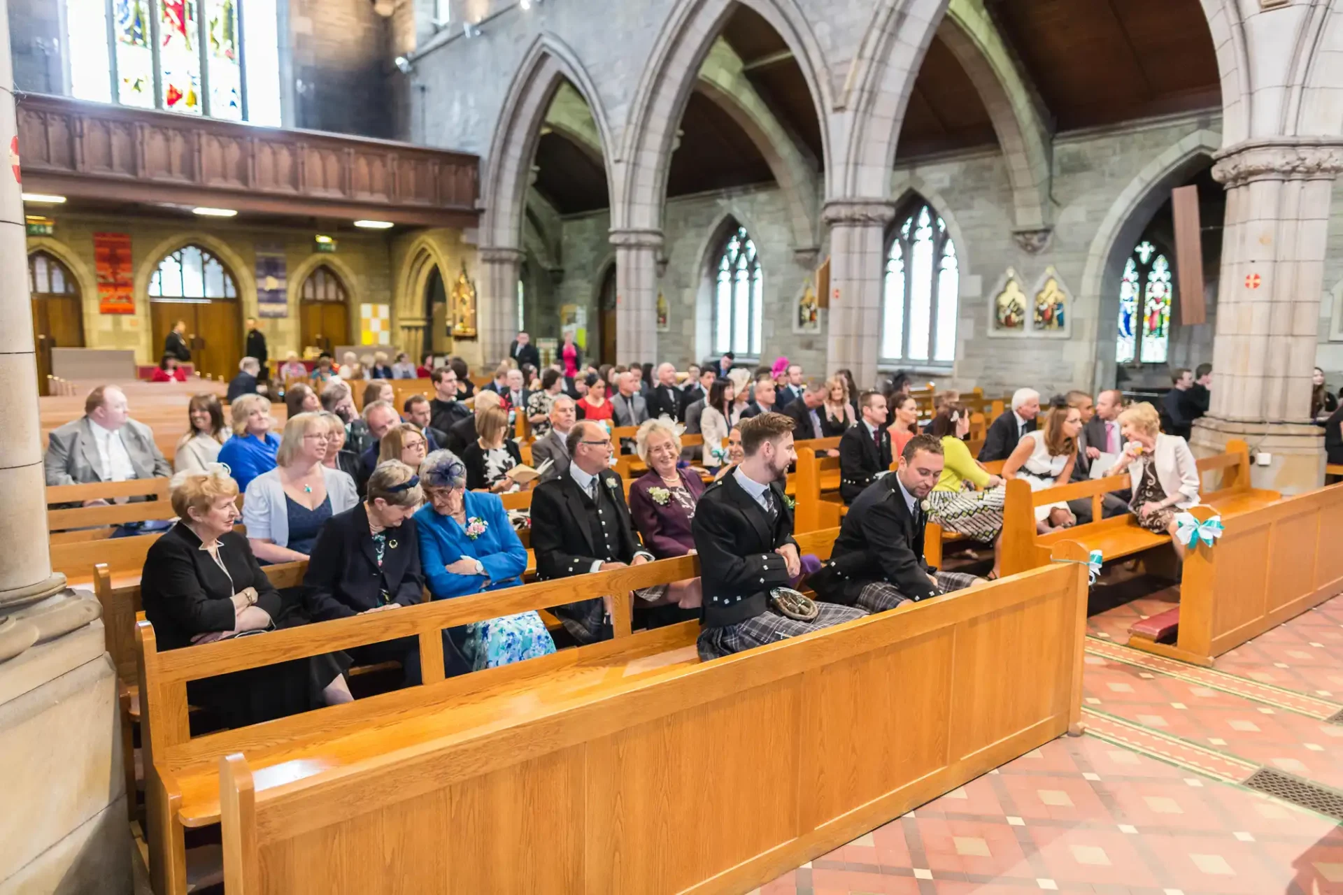 A congregation of people seated in wooden pews inside a church during a ceremony, with stained glass windows visible in the background.