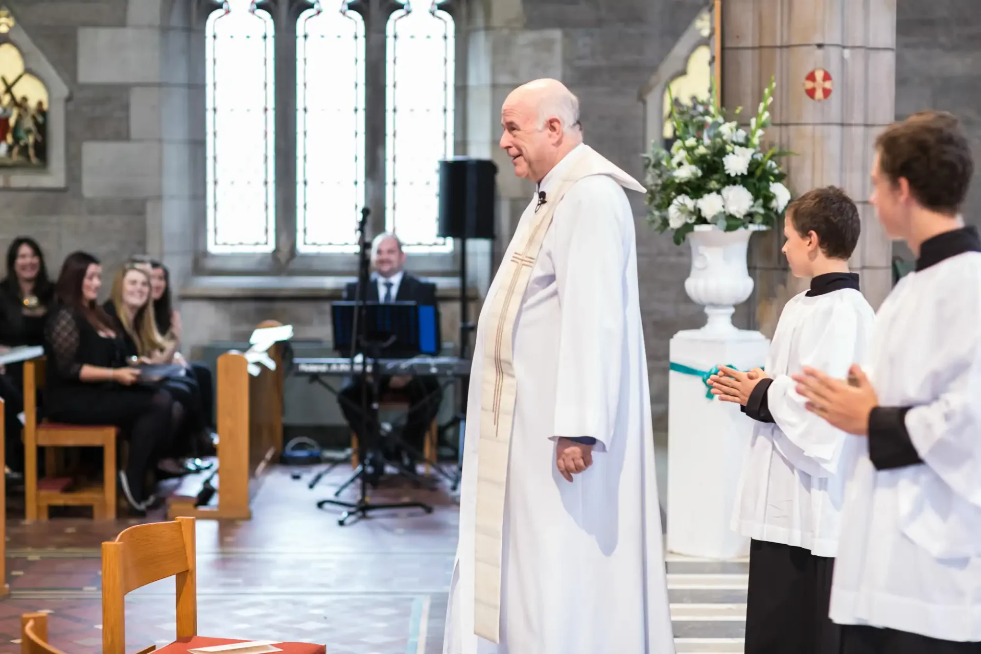 A priest speaking during a ceremony in a church with stained glass windows, attendees smiling in the background.