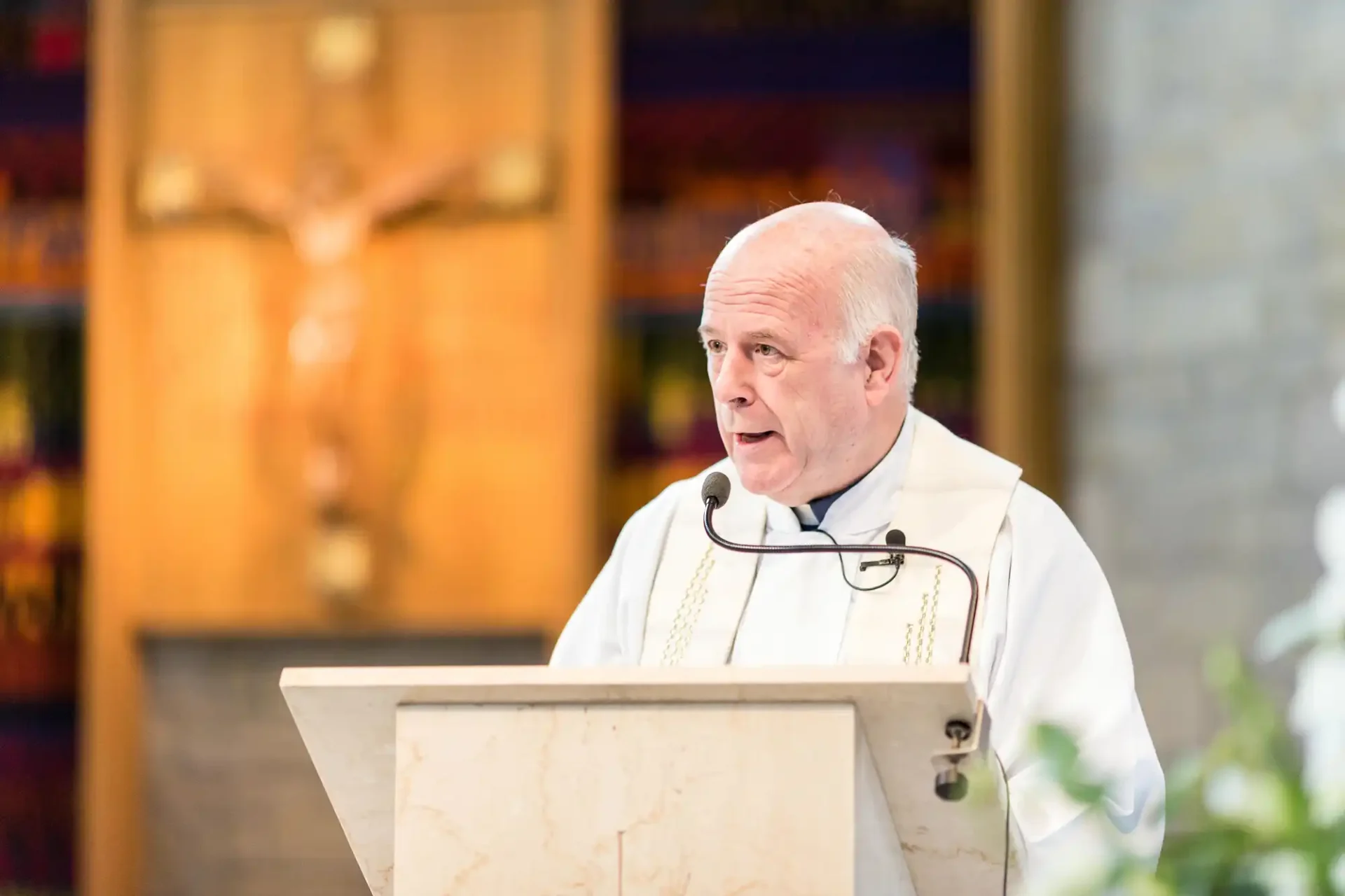 A priest speaking at a lectern in a church, with a crucifix visible in the background.