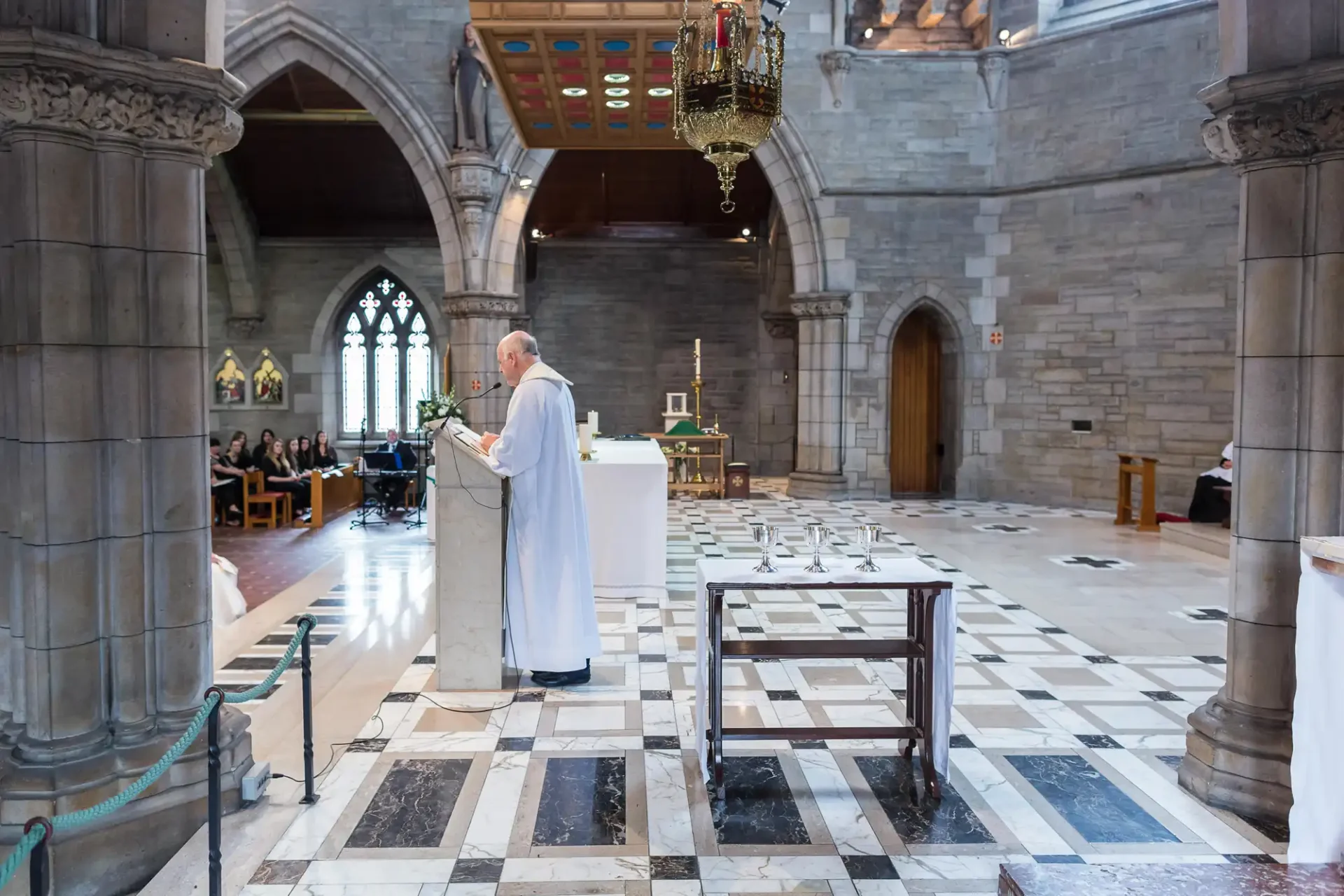 Priest delivering a sermon in a cathedral with congregants seated in the background and a lectern in the foreground.