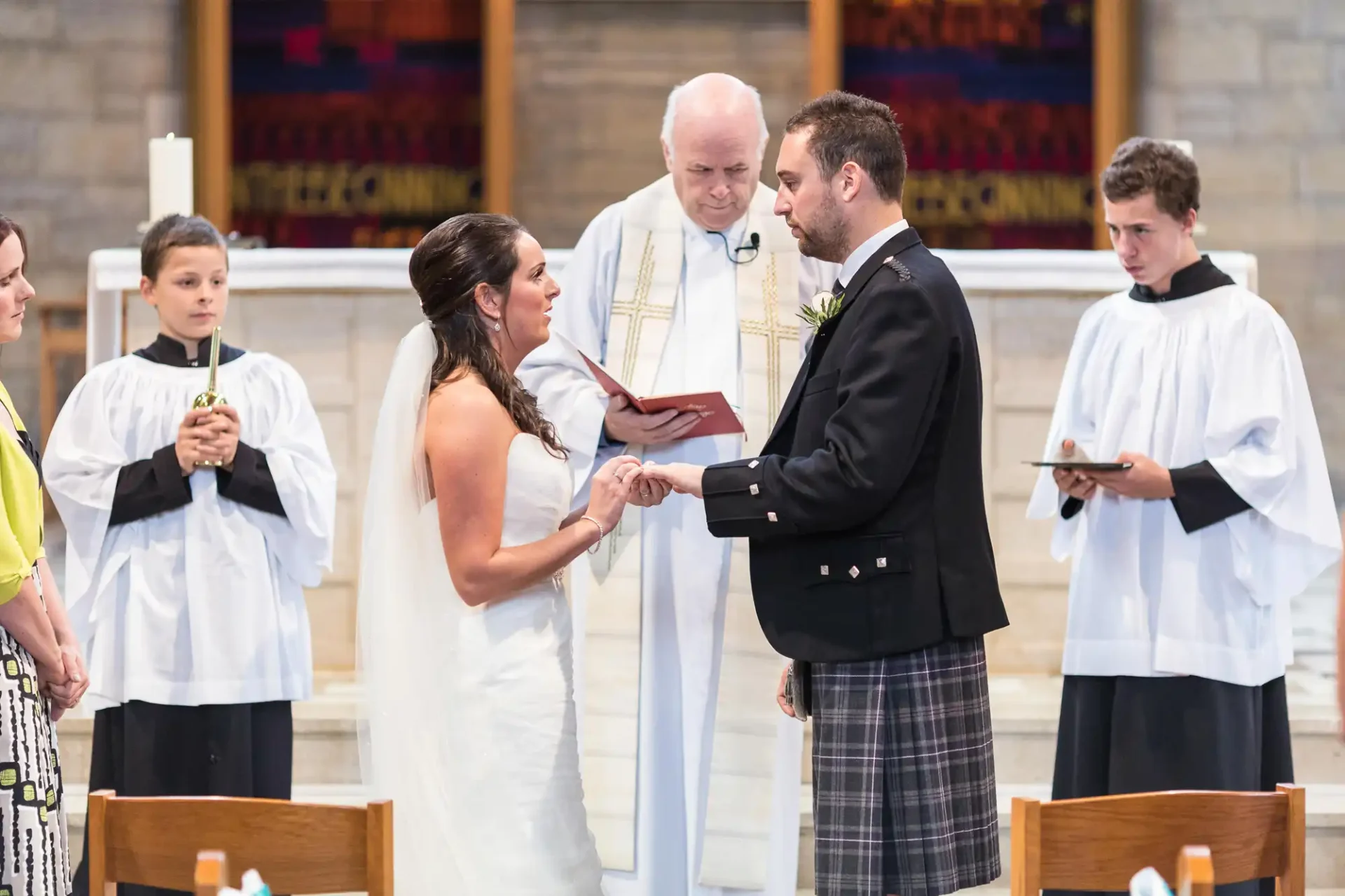 A bride and groom exchanging rings during a wedding ceremony in a church, presided over by a priest, with attendants looking on.