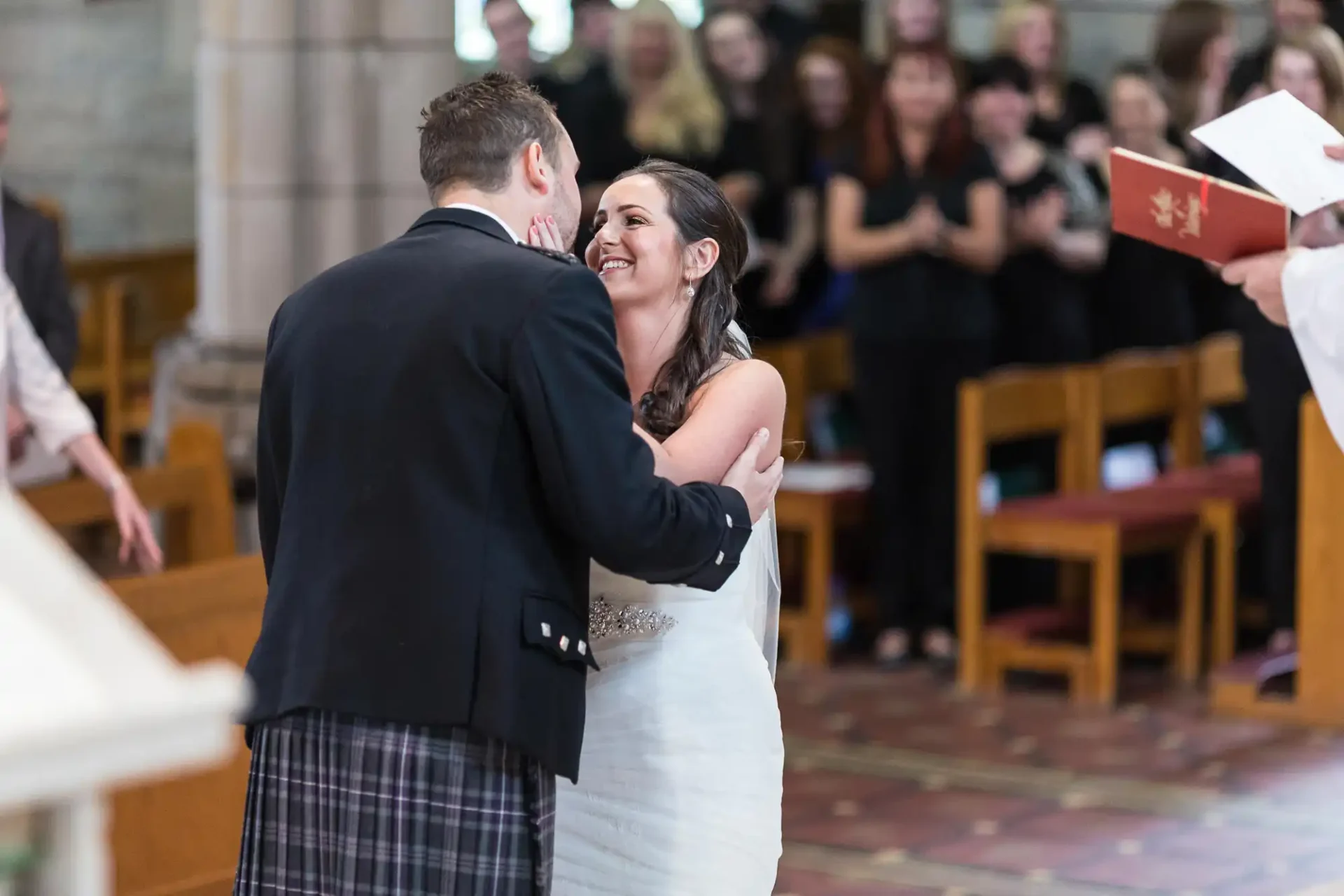 Bride and groom smiling and embracing during a church wedding ceremony, with guests in the background.