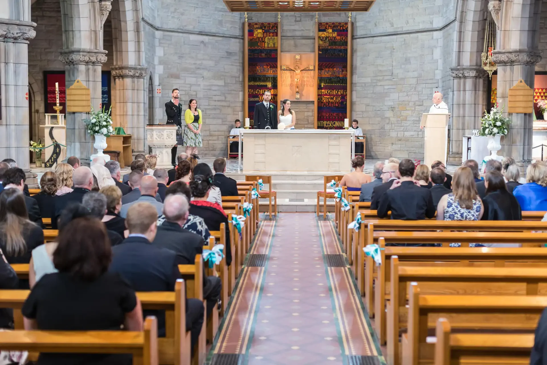 A wedding ceremony in a church with guests seated in pews looking towards a couple at the altar, flanked by floral arrangements.
