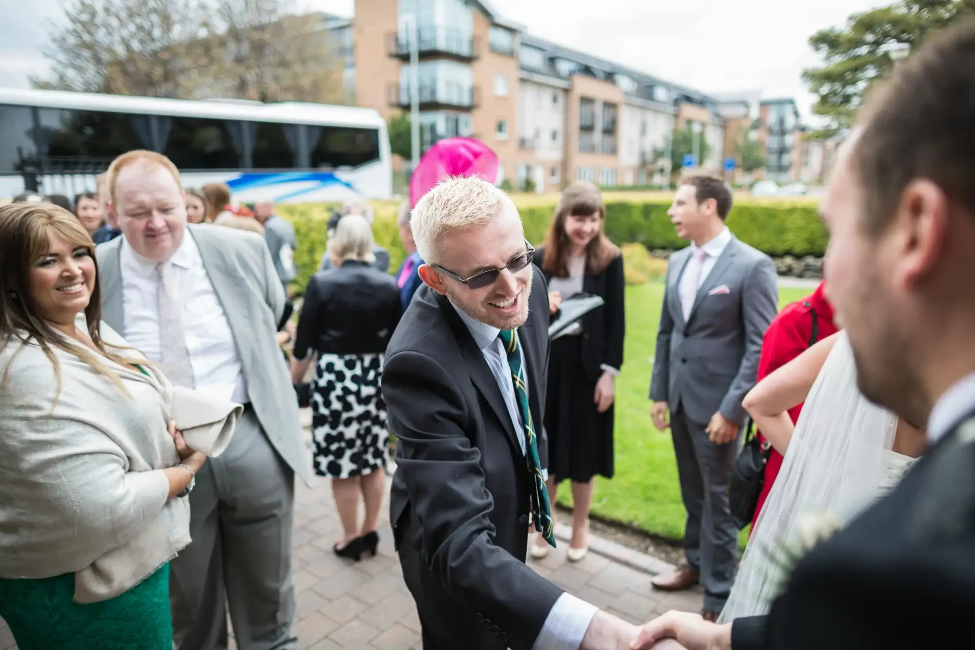 A man in a suit and sunglasses shakes hands with another man at an outdoor gathering with several people chatting in the background.