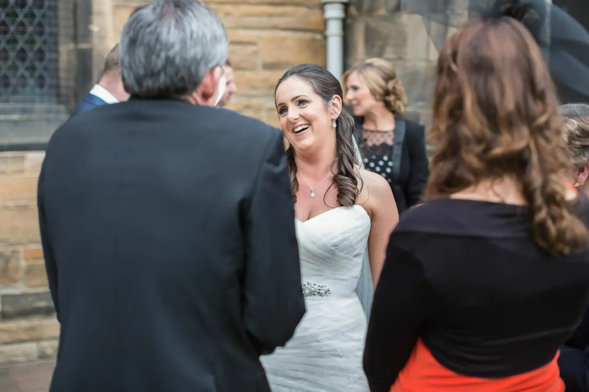 A bride in a white dress and a diamond waistband smiles at a man in a suit, with guests in the background at an outdoor event.