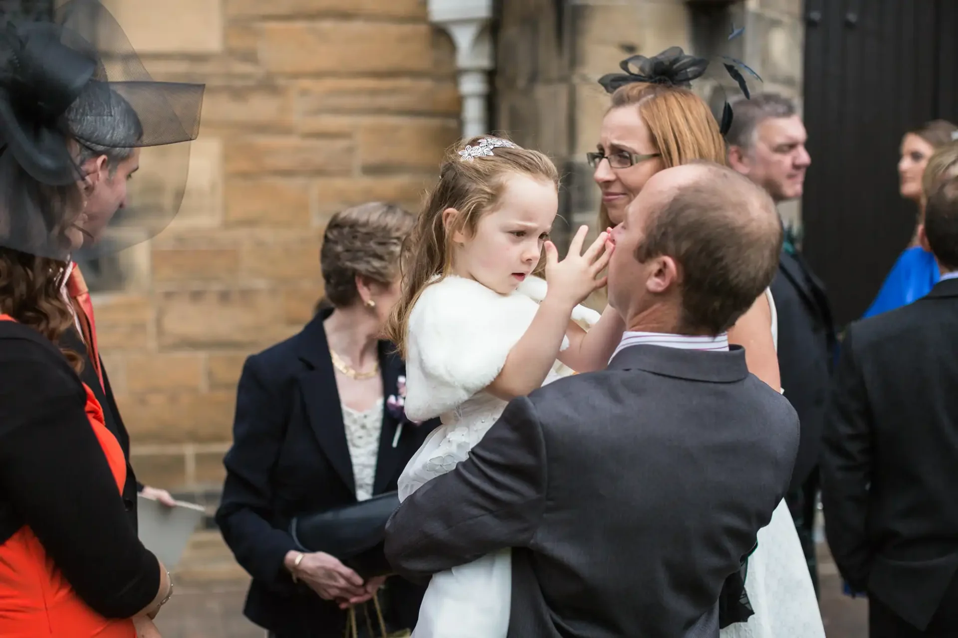 A man kneels to speak with a young girl in a white dress at a formal gathering, as a woman standing behind the girl looks on.