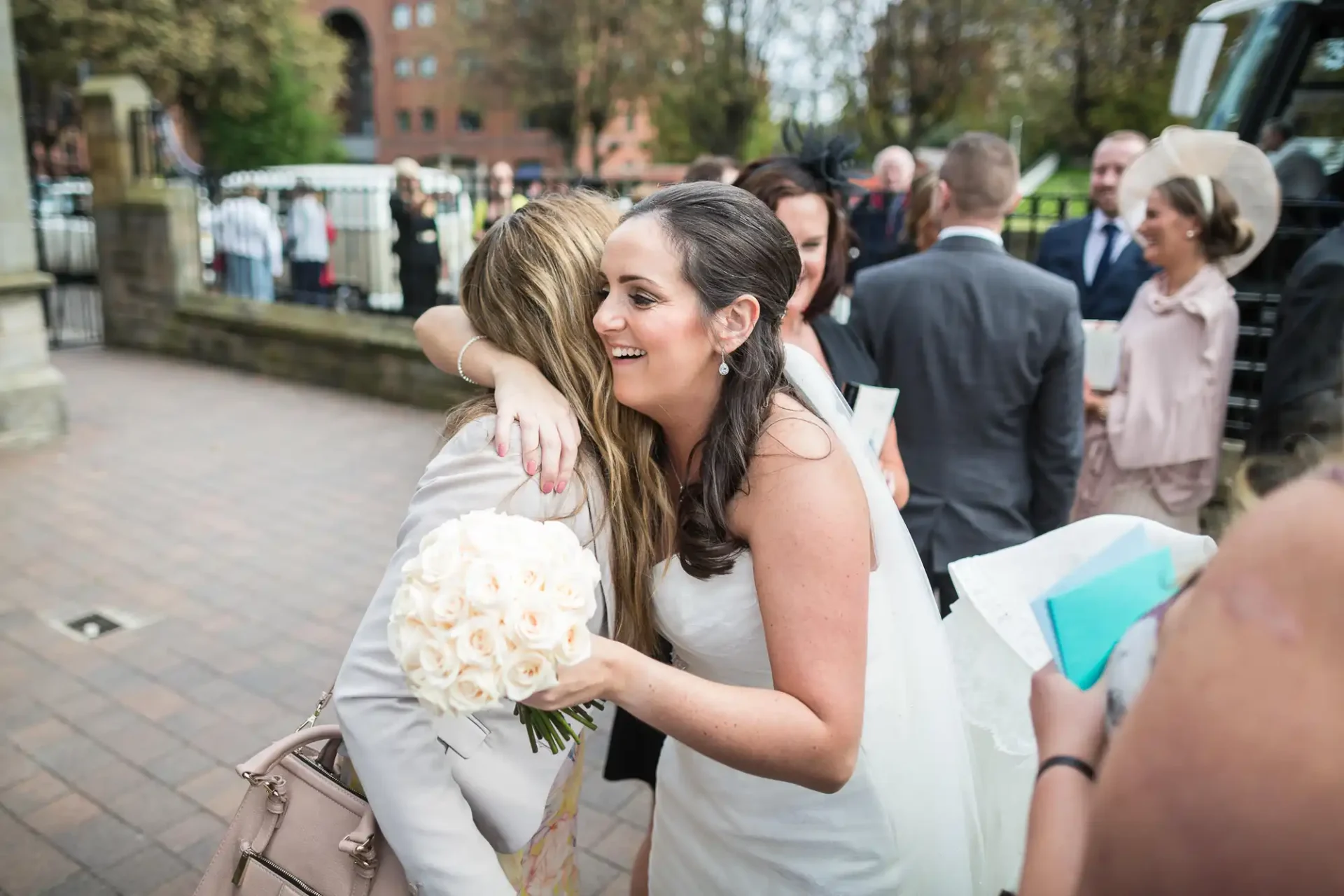Two women embracing at a wedding, one in a white bridal gown holding a bouquet, the other in a floral dress, with guests and buildings in the background.