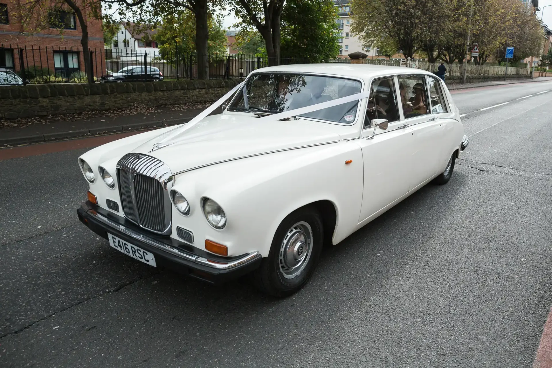 A vintage white daimler limousine parked on a city street beside a sidewalk with trees.