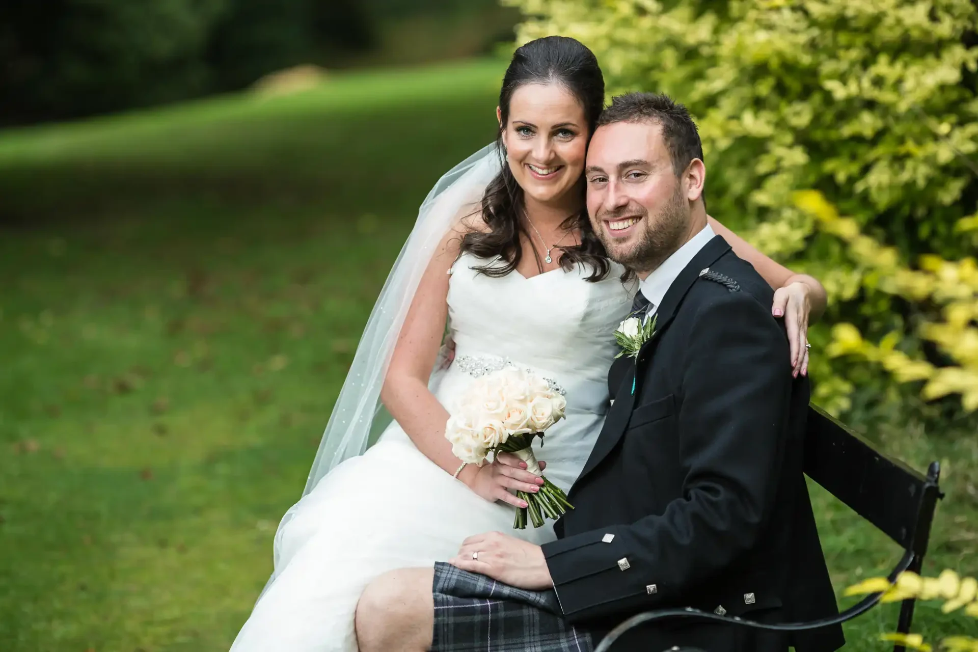 A bride and groom smiling, seated together outdoors with the bride holding a bouquet of white flowers.