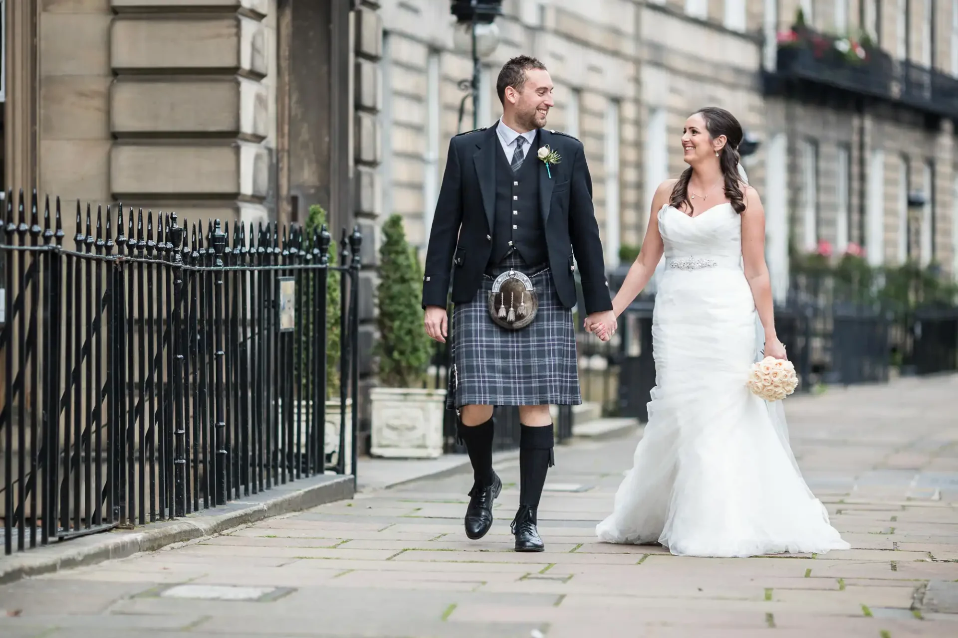 Royal Scots Club Edinburgh wedding photographer image of a newlywed couple smiling and walking down a city street, the groom wearing a kilt and the bride in a white gown.