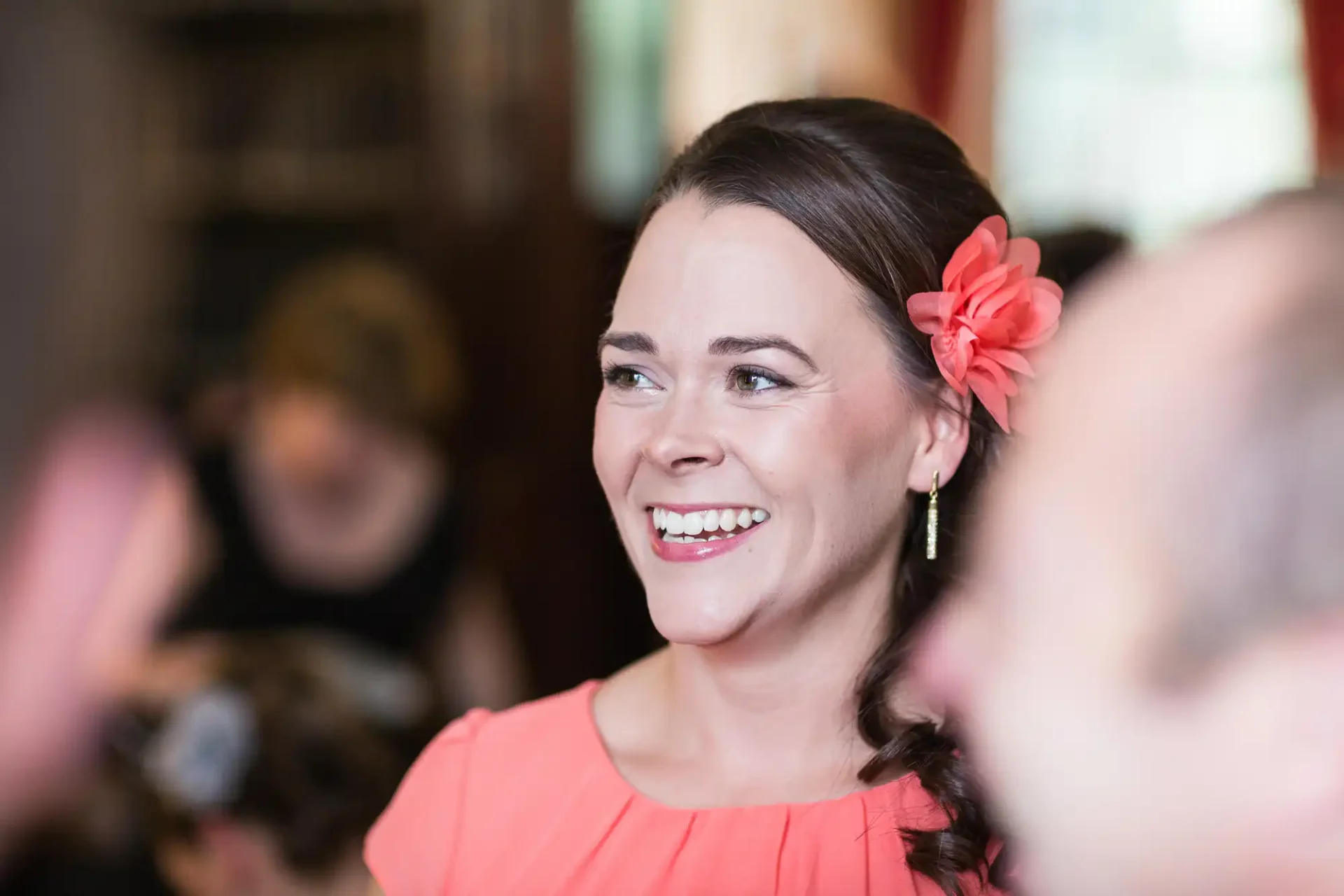 A joyful woman with a red flower in her hair and coral dress, smiling and looking away at a social gathering.