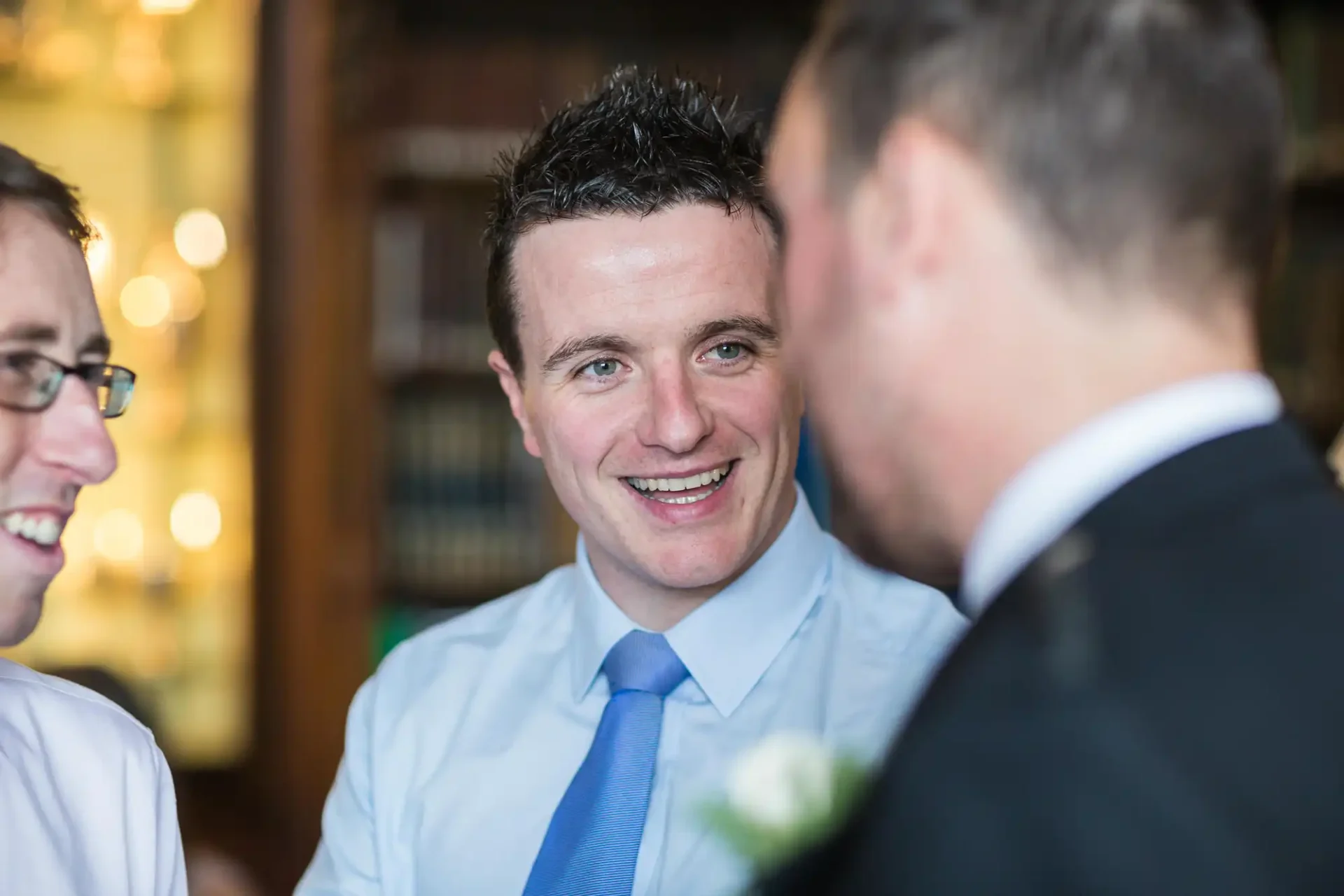 Three men engaging in conversation, with the central man smiling broadly, wearing a light blue tie and a boutonniere, in a warmly lit indoor setting.