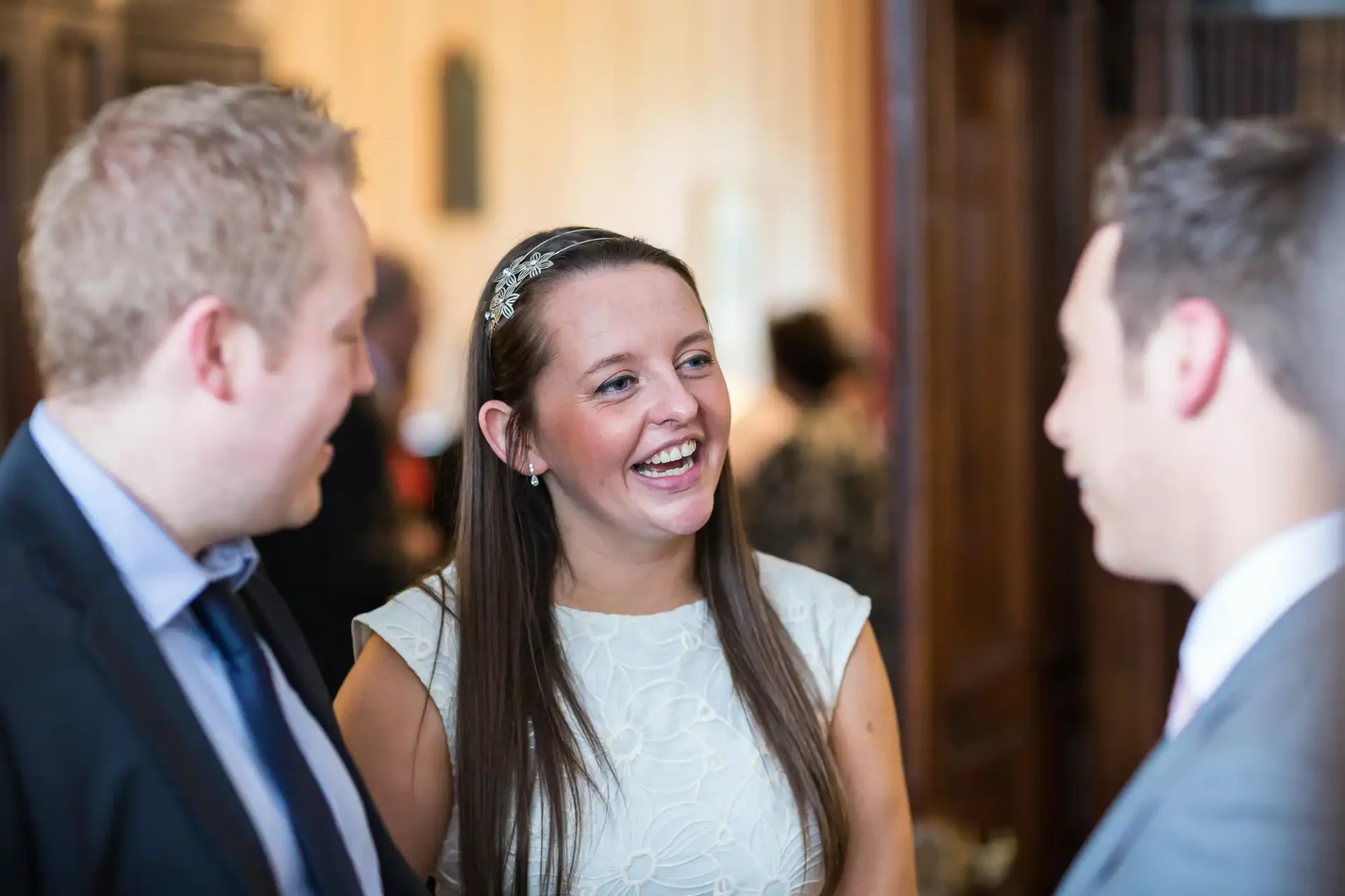 A woman in a white dress smiles while talking with two men in suits at a formal event.
