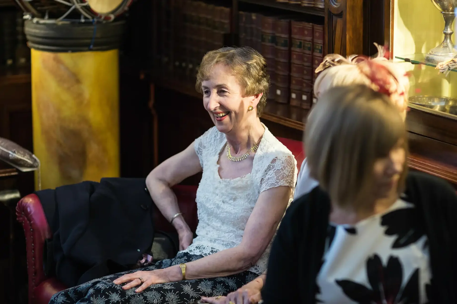 An older woman in a lace top smiles while seated on a red armchair in an elegantly furnished room, a blurred woman in the foreground.
