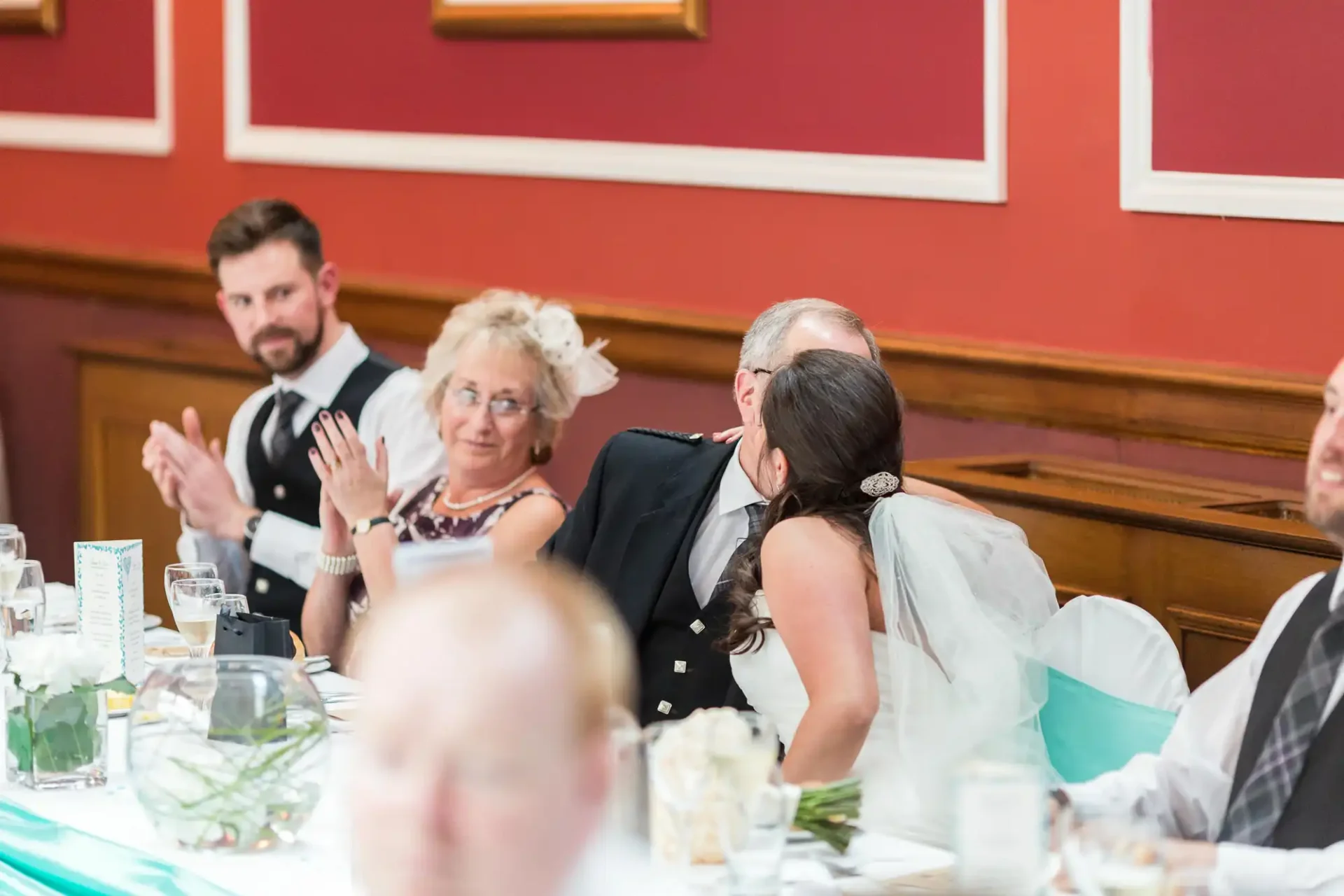 A bride in a white dress leans back to kiss an elderly man on the cheek at a wedding reception table, while a man and a woman observe, clapping.