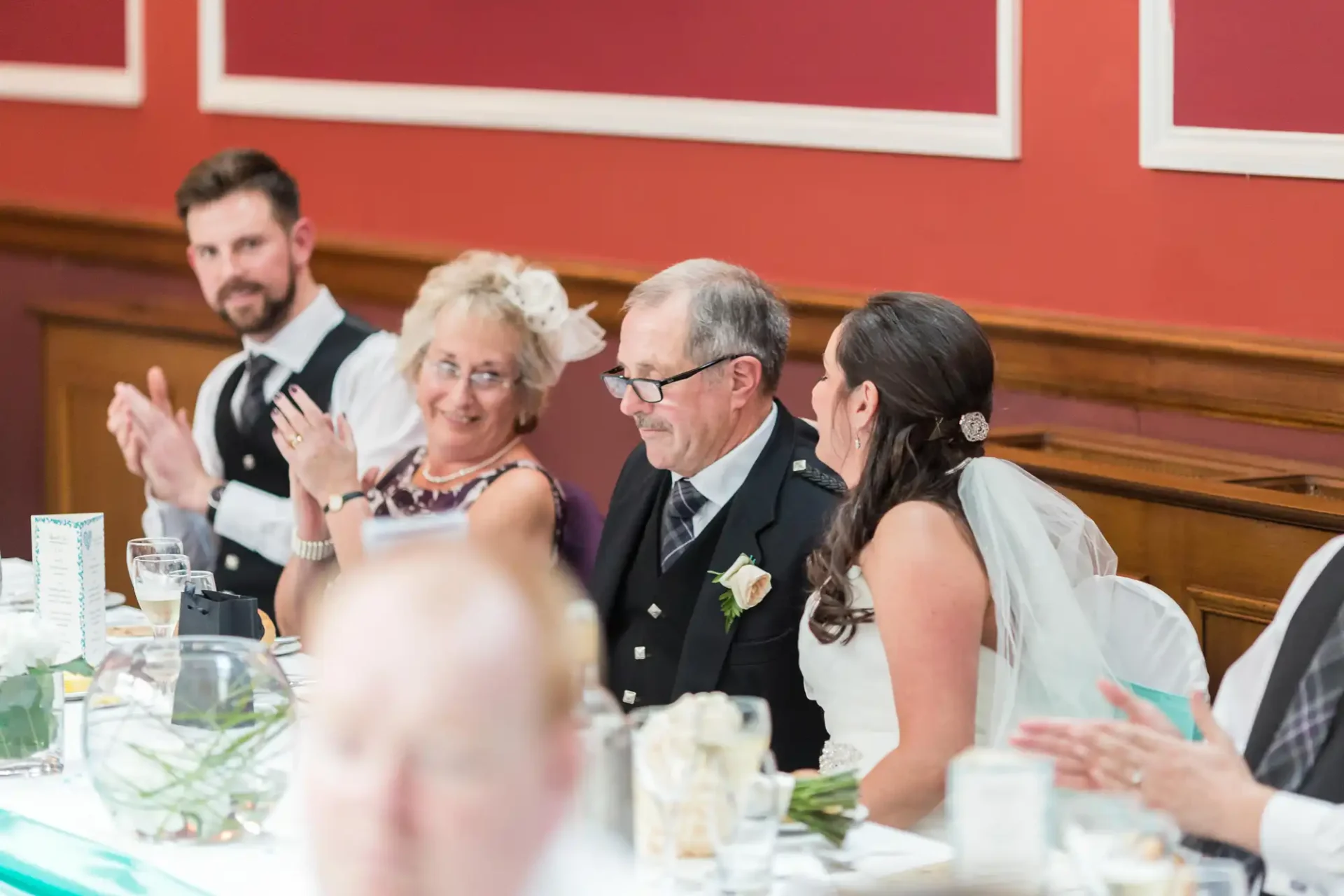 A wedding reception scene showing a bride and groom seated at a table with guests, engaging in conversation and clapping.
