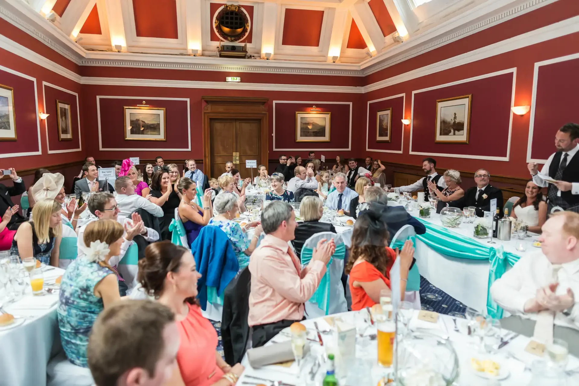 Elegant indoor wedding reception with guests seated at round tables, clapping as a speaker stands at the front of the room.