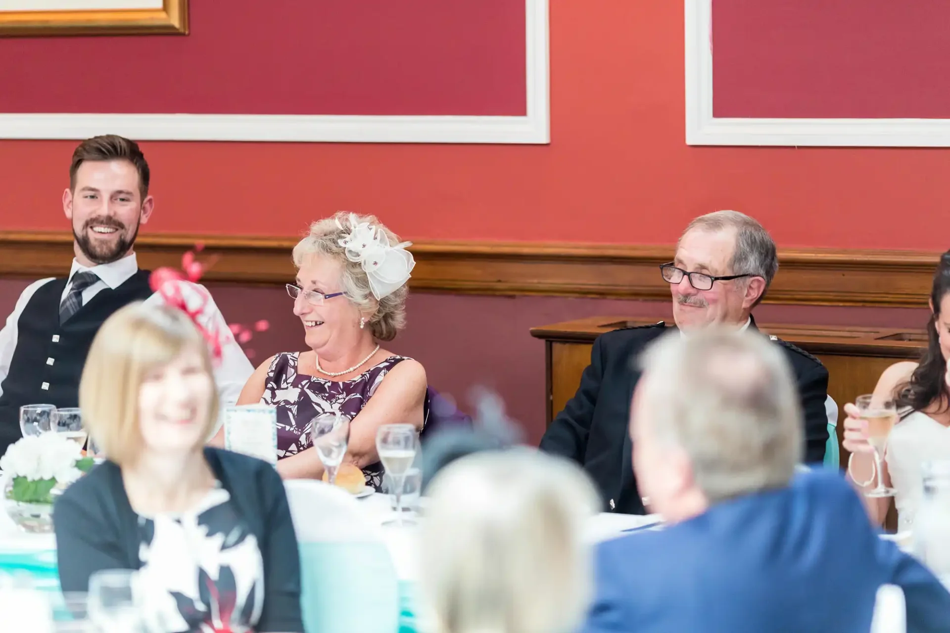 Group of elegantly dressed adults smiling and chatting at a formal dining event with red walls and white table settings.