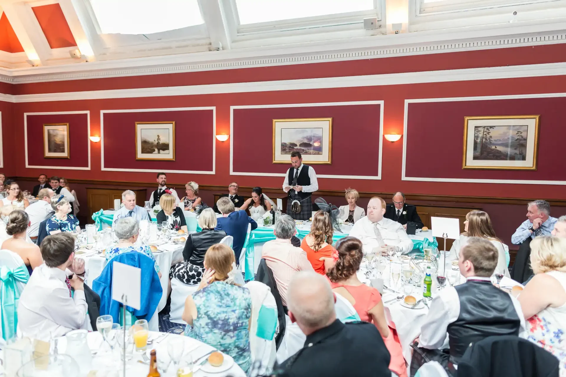 A man stands and speaks at a podium during a formal dinner event in a room with red walls and paintings, surrounded by seated guests.
