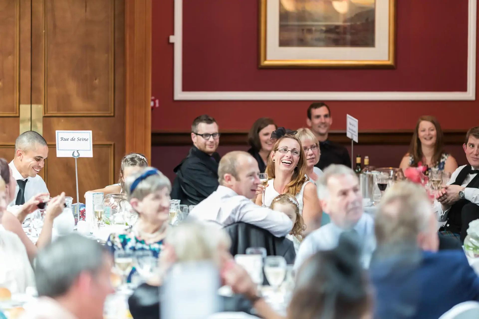 Guests seated at tables, enjoying a wedding reception in an elegant dining hall with large windows and rich wood paneling.