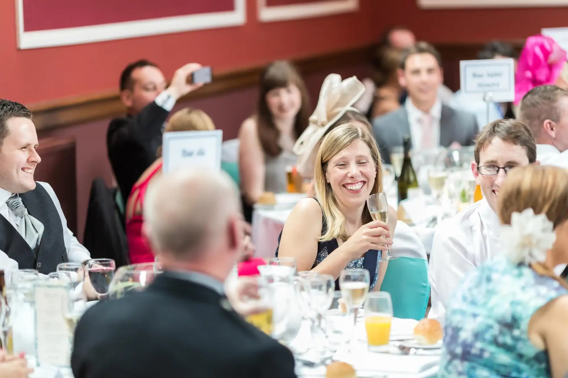 Guests at a festive event, seated at dining tables laughing and conversing, with some taking photos; focus on a smiling woman holding a glass.