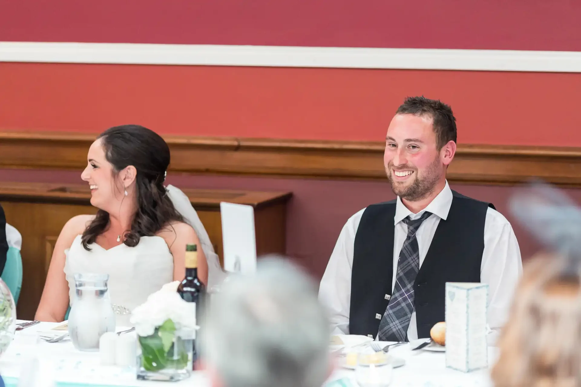 A bride and groom smiling and seated at a wedding reception table adorned with white flowers.