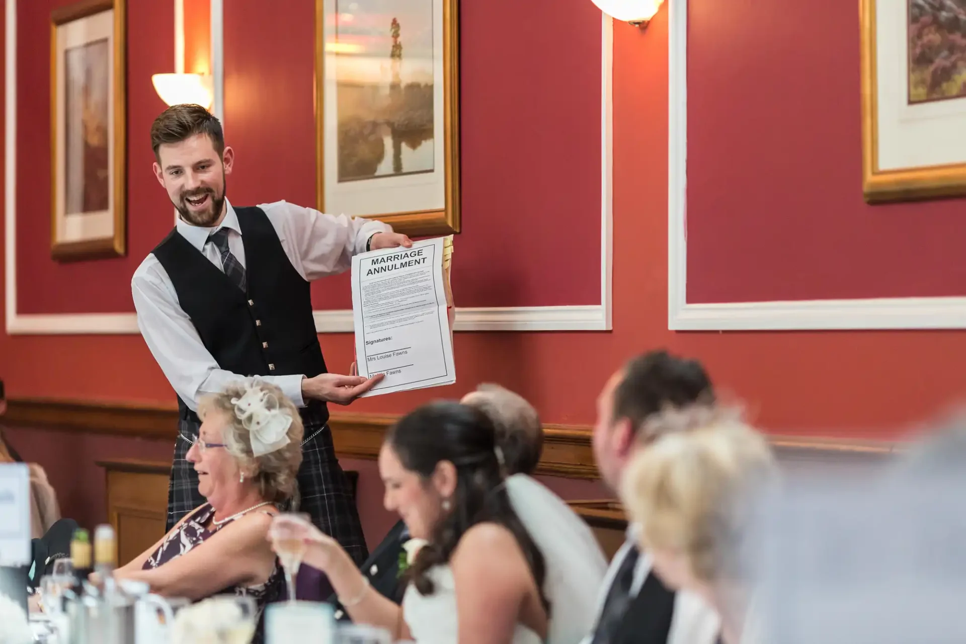 A smiling waiter presents a large certificate to guests at a banquet with paintings and people in elegant attire in the background.
