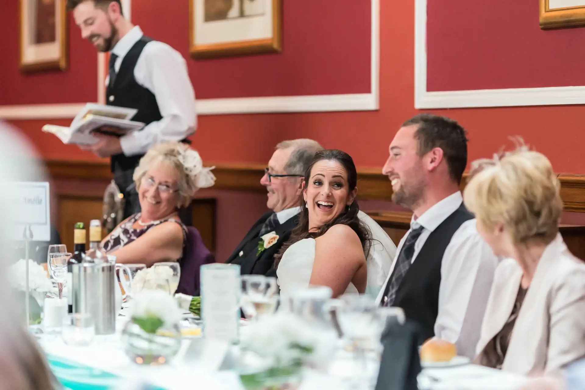 A bride smiling at the camera while seated at a table with guests during a wedding reception, with a waiter serving in the background.