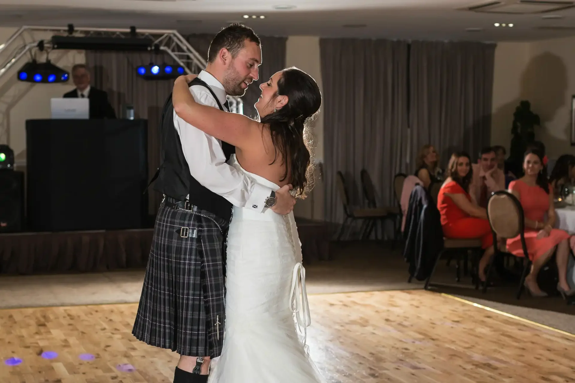 A bride and groom share a dance at their wedding reception, surrounded by seated guests, with a dj booth in the background.