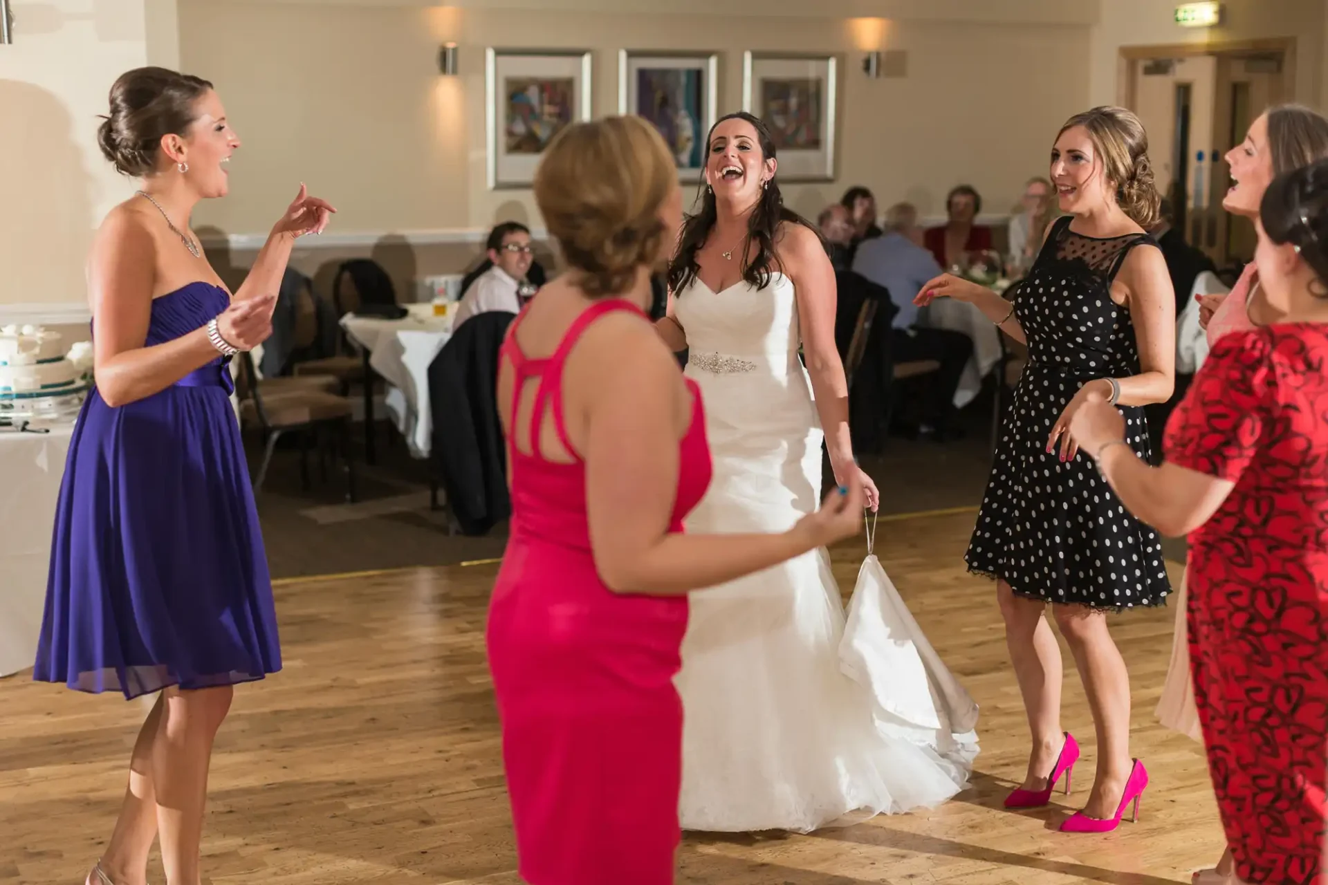 A bride in a white dress laughing and dancing with four women at a wedding reception, with guests seated in the background.