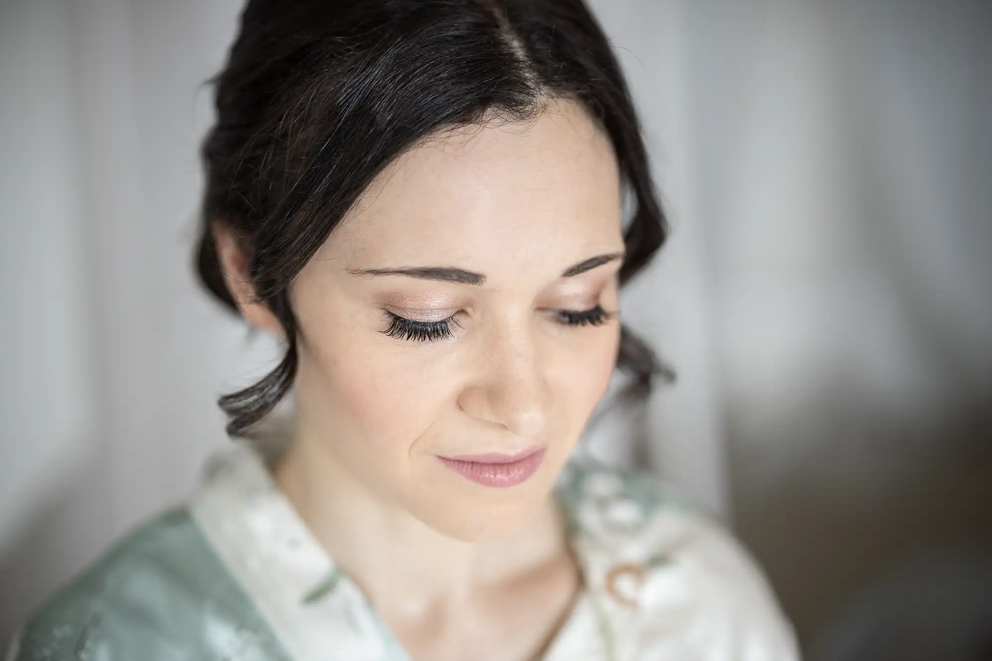 A woman with dark hair and closed eyes is looking downward, wearing a light-colored garment, set against a soft, neutral background.