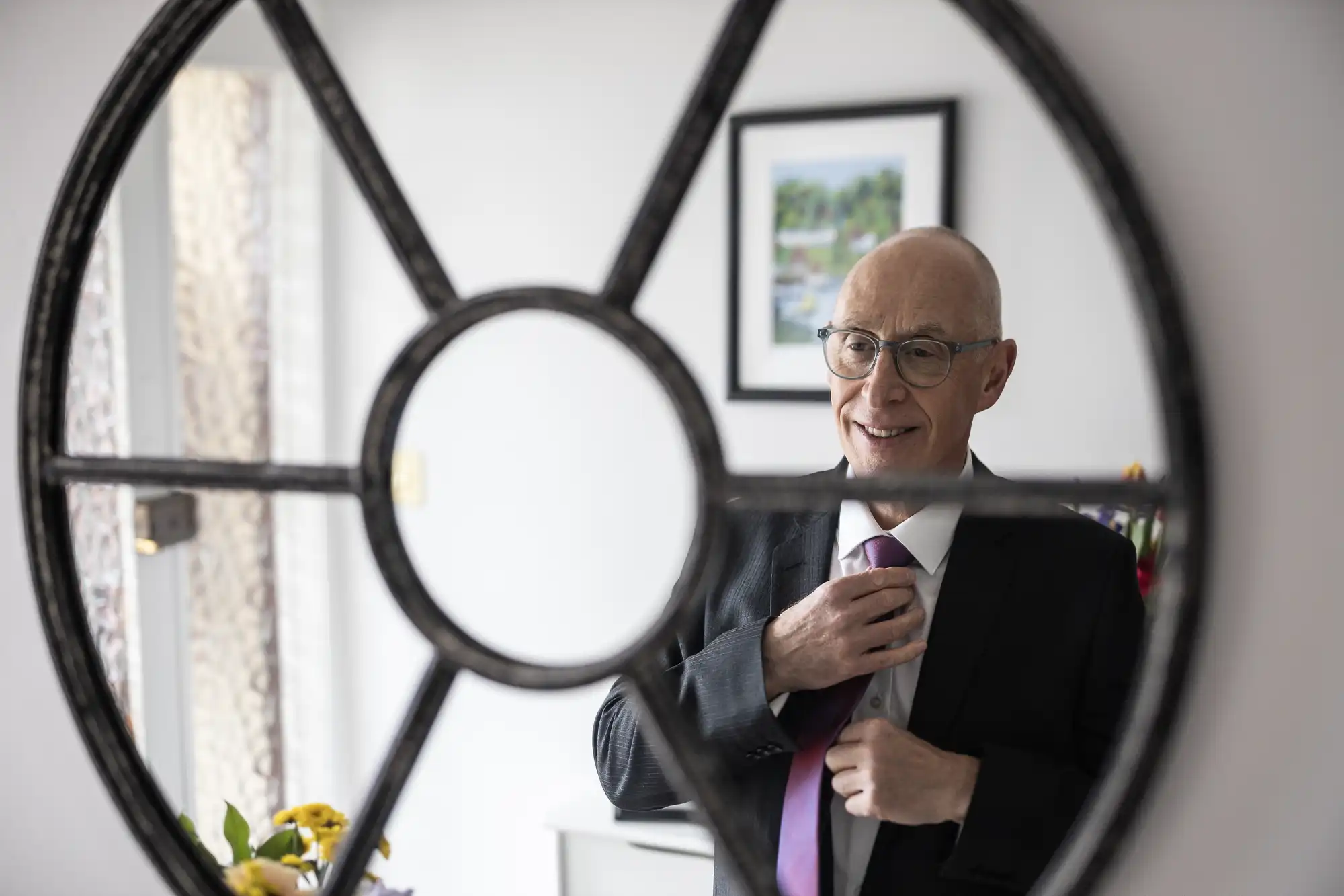 A man in a suit adjusts his tie, reflected in a round mirror with a decorative frame, in a room with a framed picture in the background.